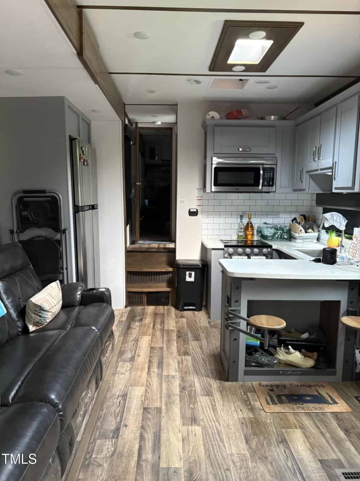 Interior of an RV with wood-look flooring, black recliner sofa, gray kitchen cabinets, white subway tile backsplash, microwave, fridge, and a small island with two stools. Steps lead to a raised area at the back.