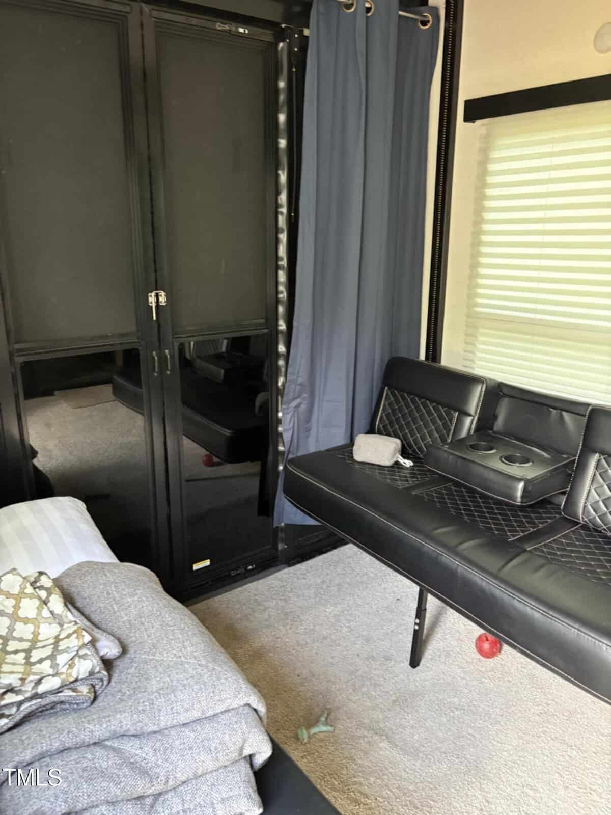 A small room with carpeted floor, a black futon with a cushion and a fabric case, a pile of blankets on a bed, mirrored closet doors, a curtain, and a window with blinds; a red ball is on the floor.
