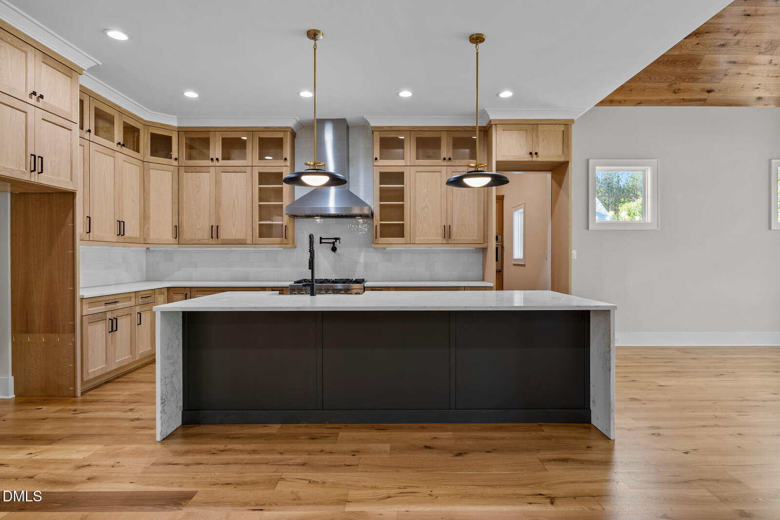 A modern kitchen with light wood cabinets, a large gray and white marble island, stainless steel appliances, two black pendant lights, and wood floors. White walls and a wood-paneled ceiling accent are also visible.