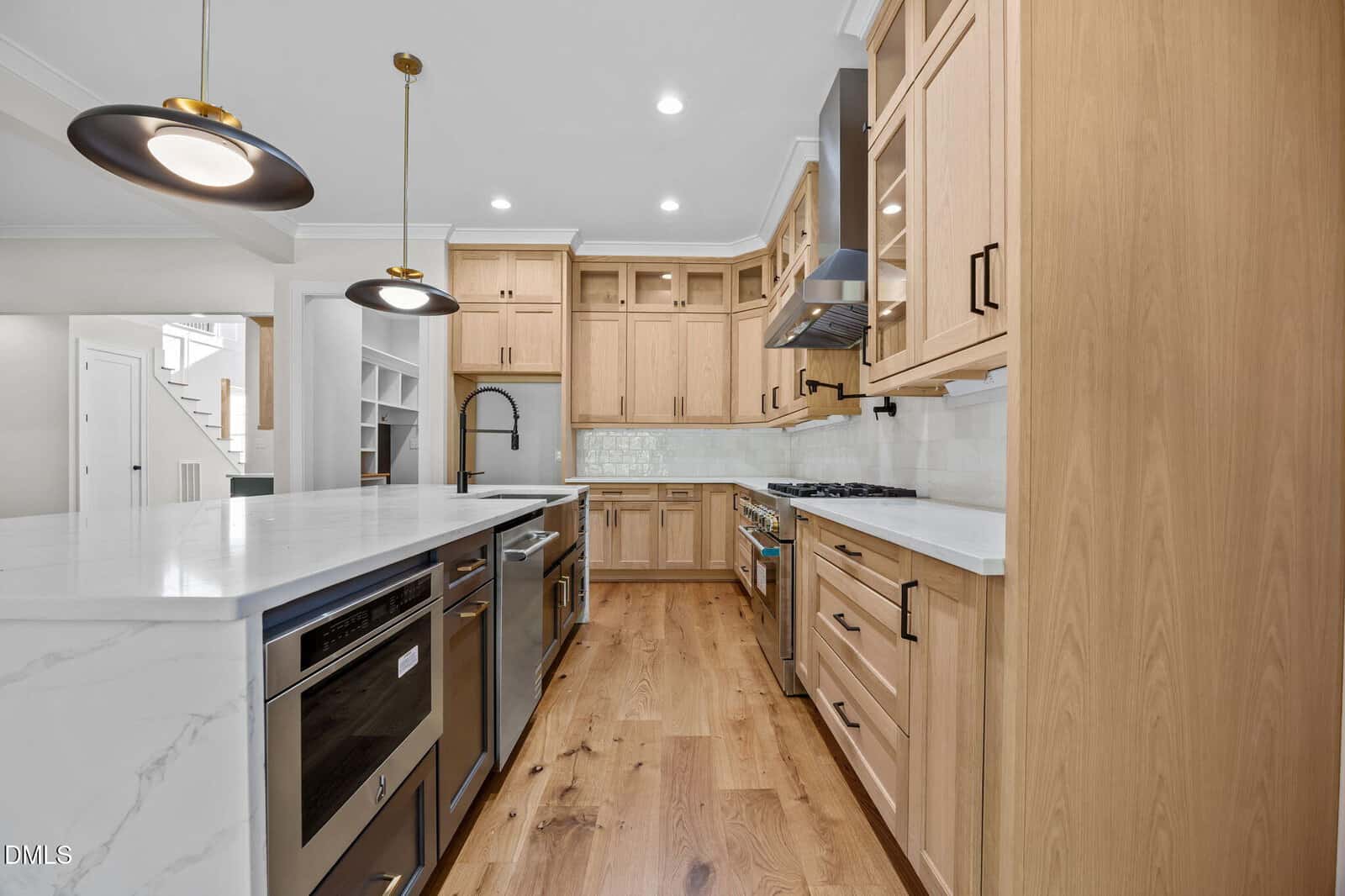 Modern kitchen with light wood cabinets, white marble countertops, stainless steel appliances, built-in stove, pendant lights, and a farmhouse sink. Hardwood floors and open shelving enhance the bright, spacious design.