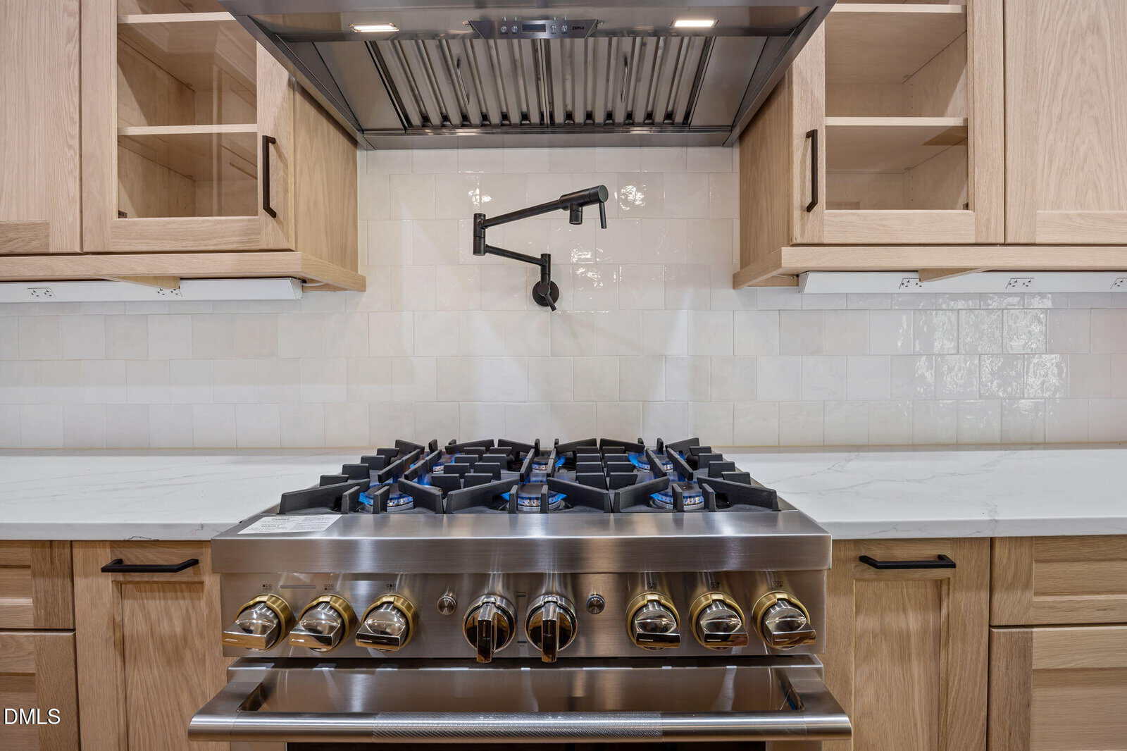 A modern kitchen stove with six burners, brass knobs, a stainless steel backsplash, light wood cabinets, white countertops, and a black pot filler faucet mounted on a white tile backsplash.