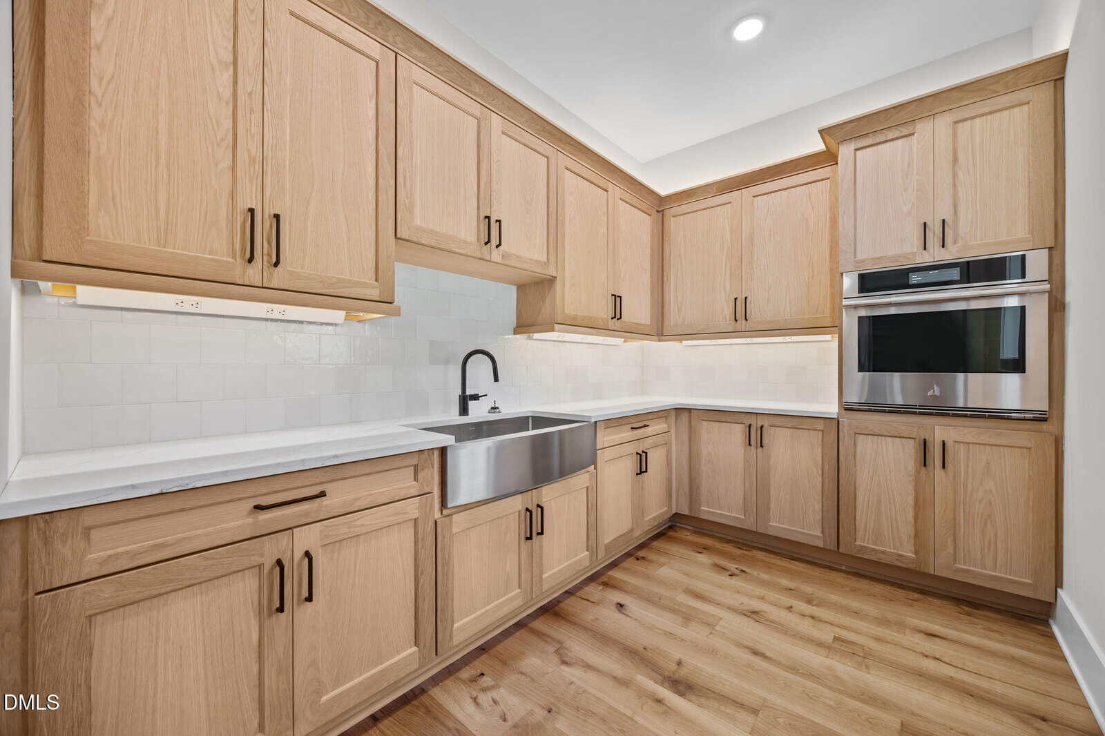 Modern kitchen with light wood cabinets, stainless steel farmhouse sink, built-in double oven, white tile backsplash, and wood flooring. Bright lighting enhances the clean and spacious appearance.