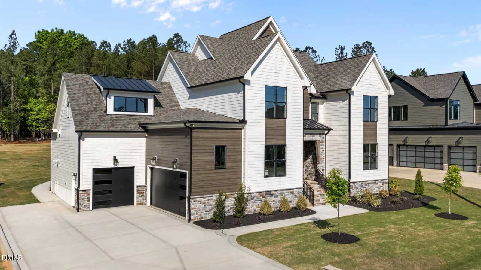 Modern two-story house with white and brown siding, multiple roof peaks, large black-trimmed windows, and a three-car garage. The front yard features young trees and neatly landscaped bushes.