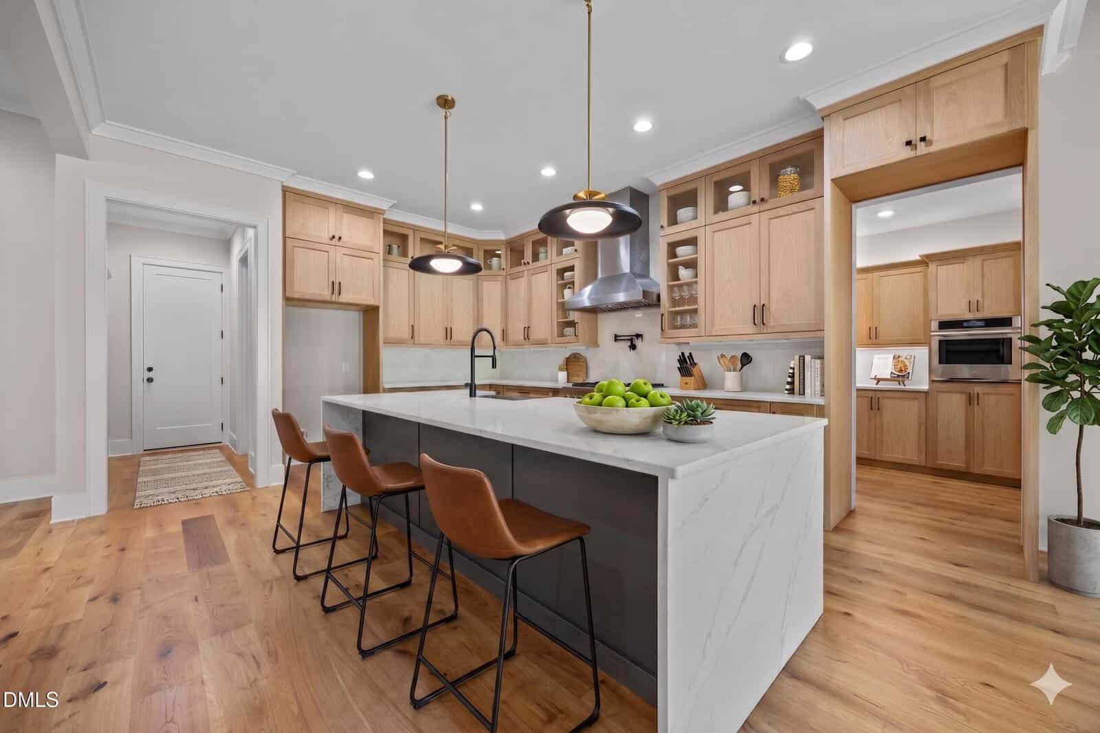 Modern kitchen with light wood cabinets, a large white marble island with sink and three brown leather barstools, pendant lights overhead, open shelves, and stainless steel appliances. Bowl of green apples on island.