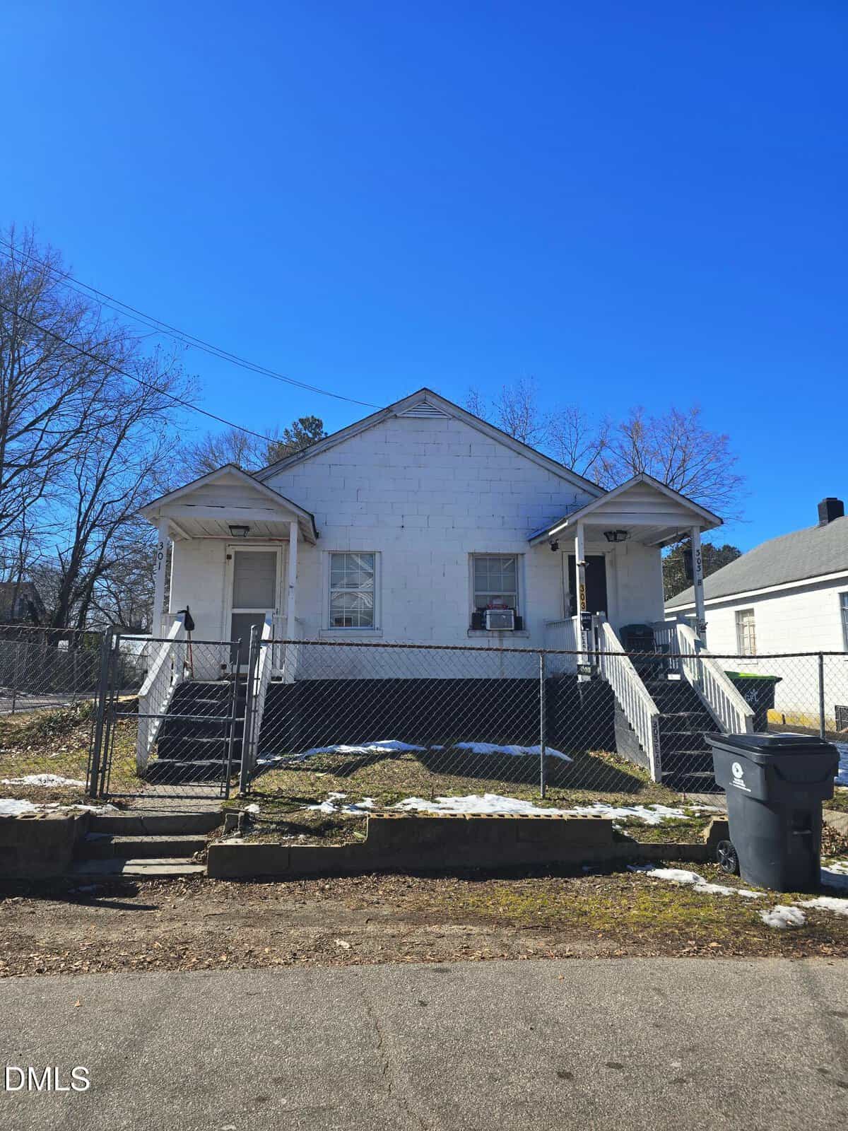 A small white duplex at 301 Pearl Street in Henderson features two front doors, each accessed by separate steps. A chain-link fence surrounds the yard, with snow patches visible on the ground. A trash bin sits by the curb, while trees and a neighboring house are nearby.