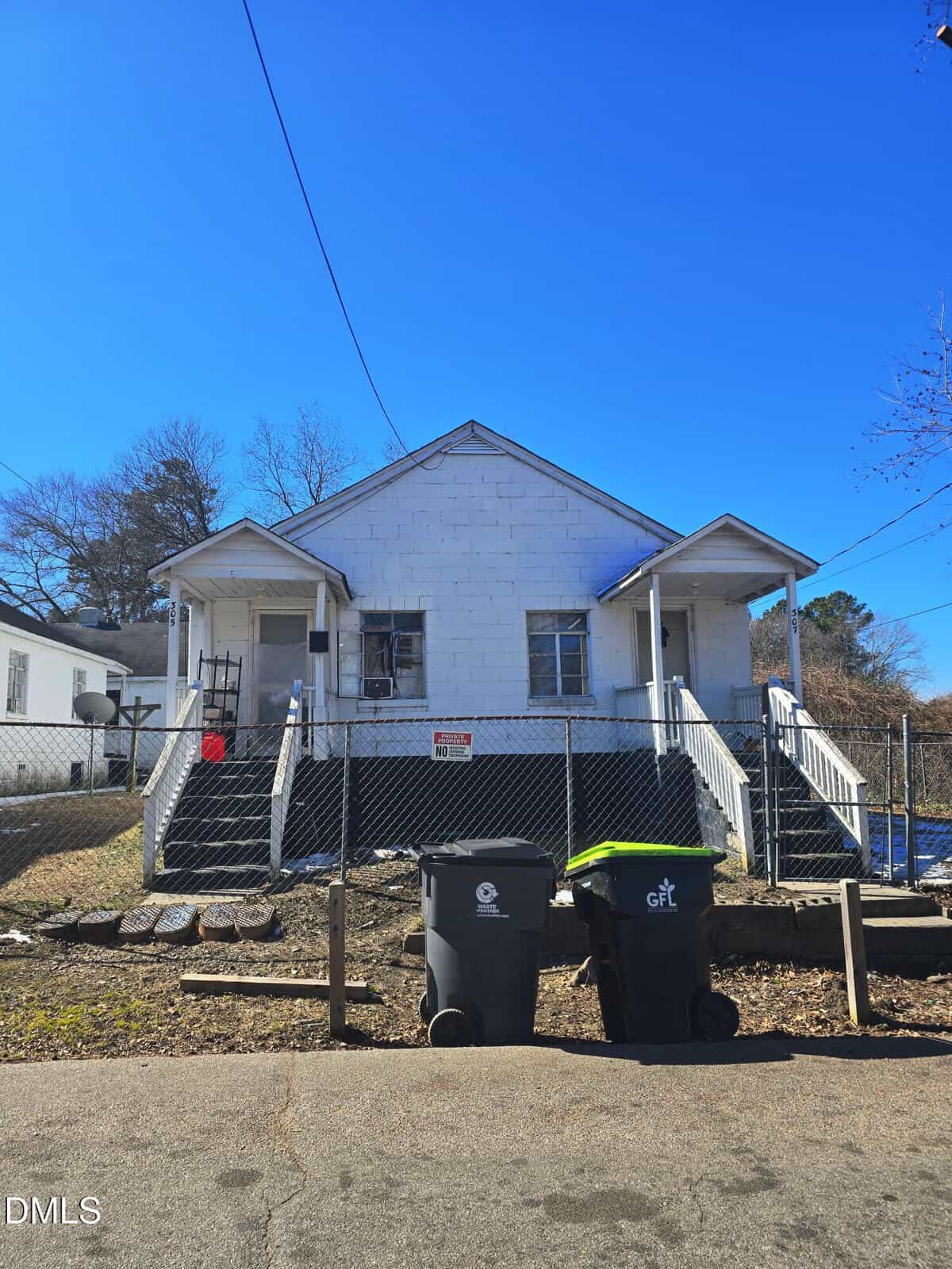 A white duplex house with two entrances, each with steps and railings, sits behind a black metal fence. Two trash bins are on the sidewalk in front. The sky is clear and blue, with leafless trees in the background.