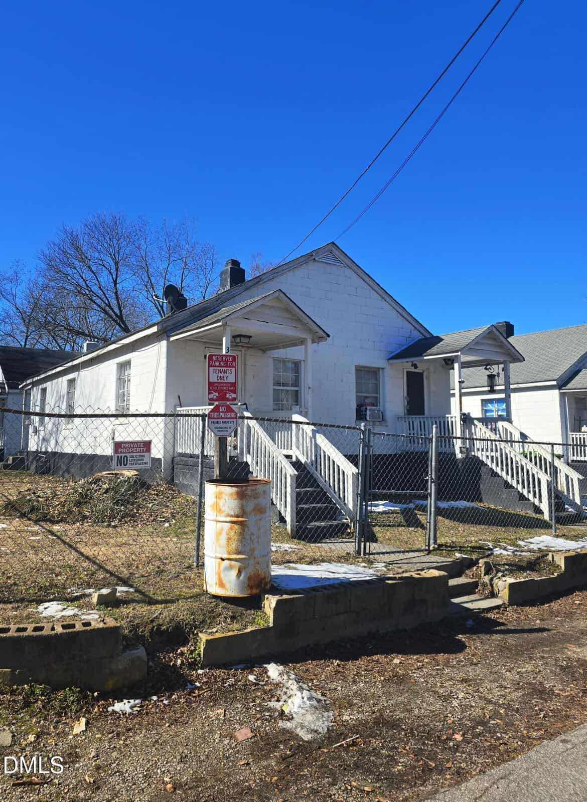 A small white house with a chain-link fence, a weathered barrel, and a “No Parking” sign out front. The yard has patches of snow, and the sky is clear and blue.