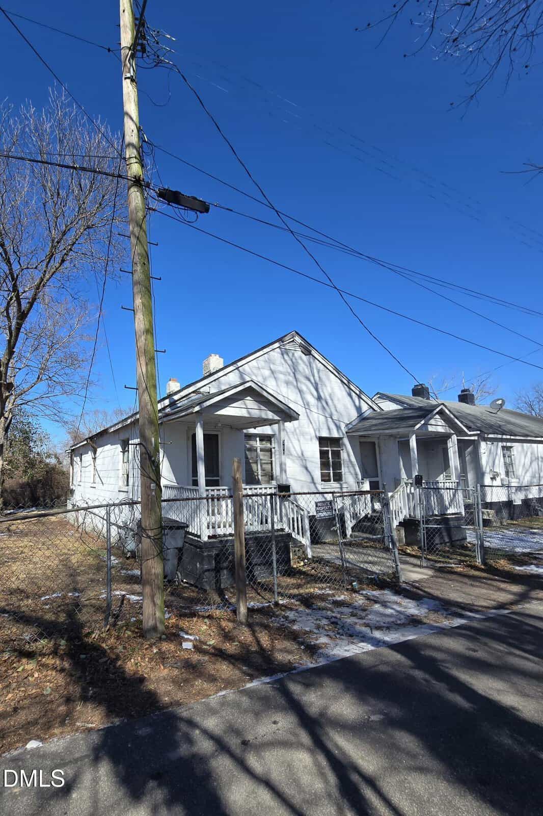 A white, single-story house with a front porch and ramp, surrounded by a chain-link fence, sits under a clear blue sky with some snow patches on the ground.