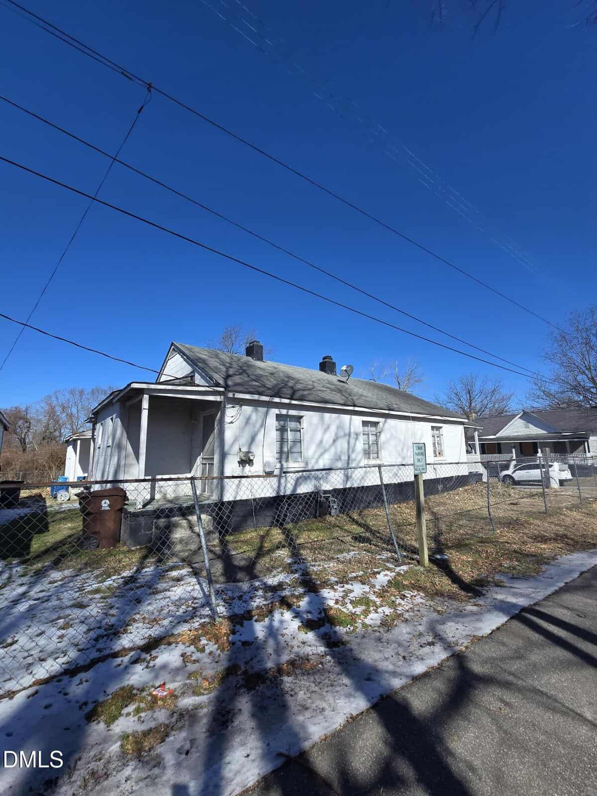 A small white house with a front porch and a fenced yard sits under a clear blue sky. There is some snow on the ground and bare trees in the background. Power lines stretch across the top of the image.