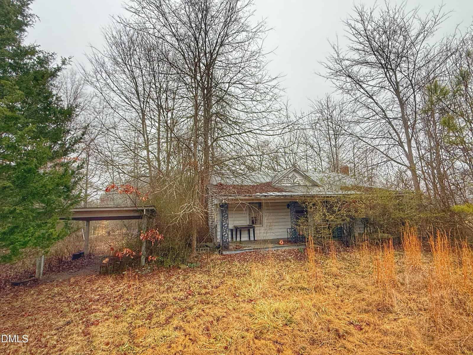 A small, weathered house sits in an overgrown yard with dry grass and leafless trees. There is a covered carport on the left side, and the house exterior appears neglected and partially obscured by bushes.