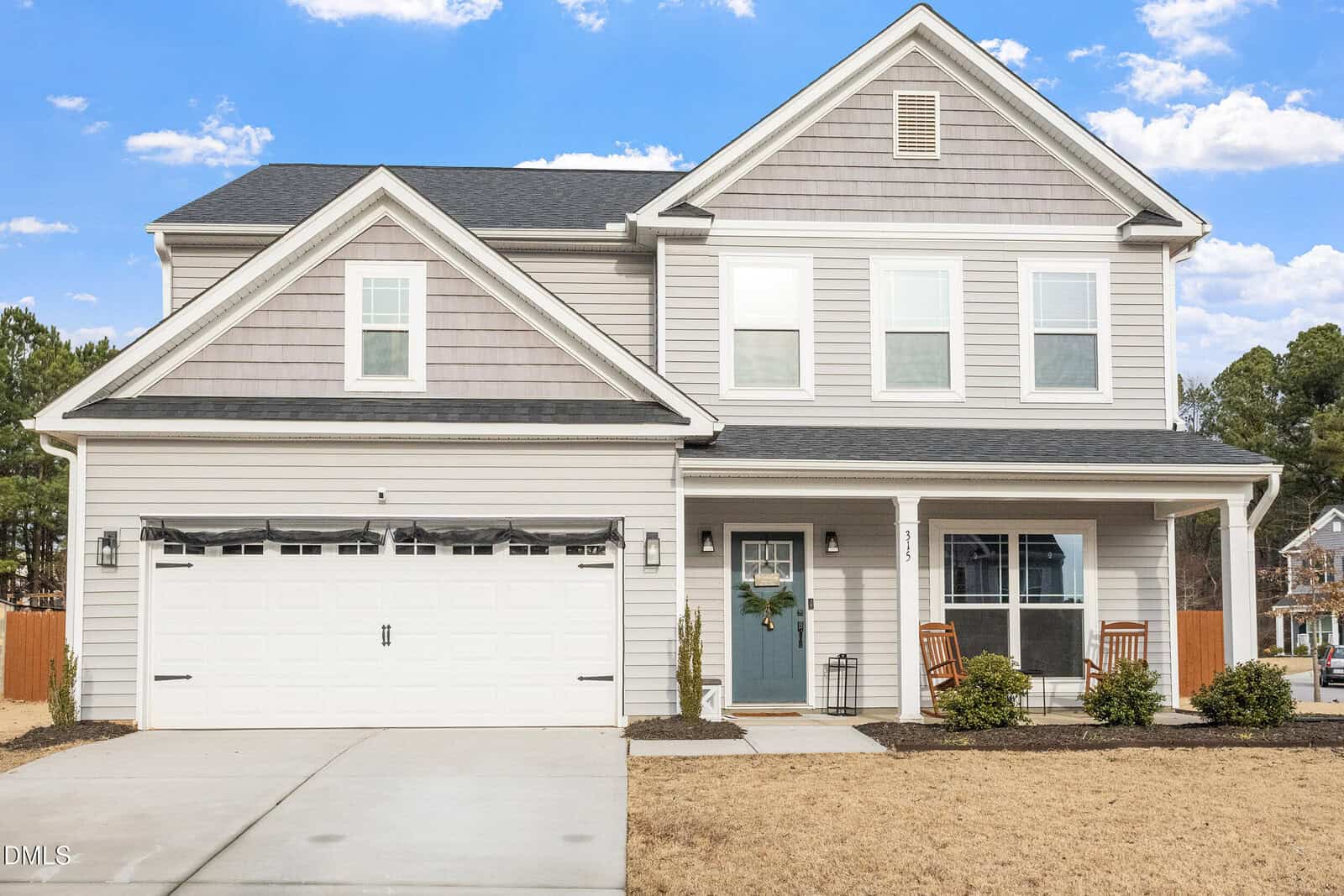 Charming two-story suburban house at 315 Shore Pine Drive, Youngsville, featuring gray siding, a double garage, small front porch with two chairs, and a blue front door. The driveway and patches of grass lead to inviting outdoor spaces, framed by trees and a bright blue sky in the background.