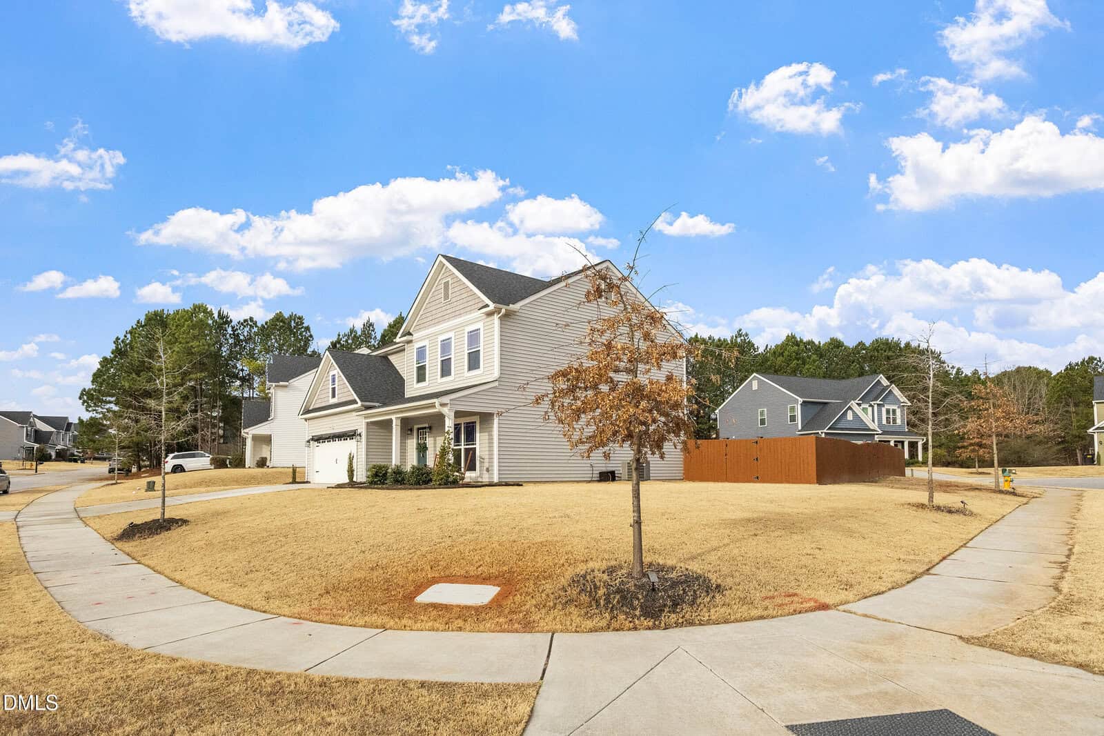 A two-story suburban house with beige siding, white trim, and a dark roof sits on a corner lot with yellowed grass and a few small trees. The neighborhood features wide sidewalks and similar houses in the background under a blue sky.