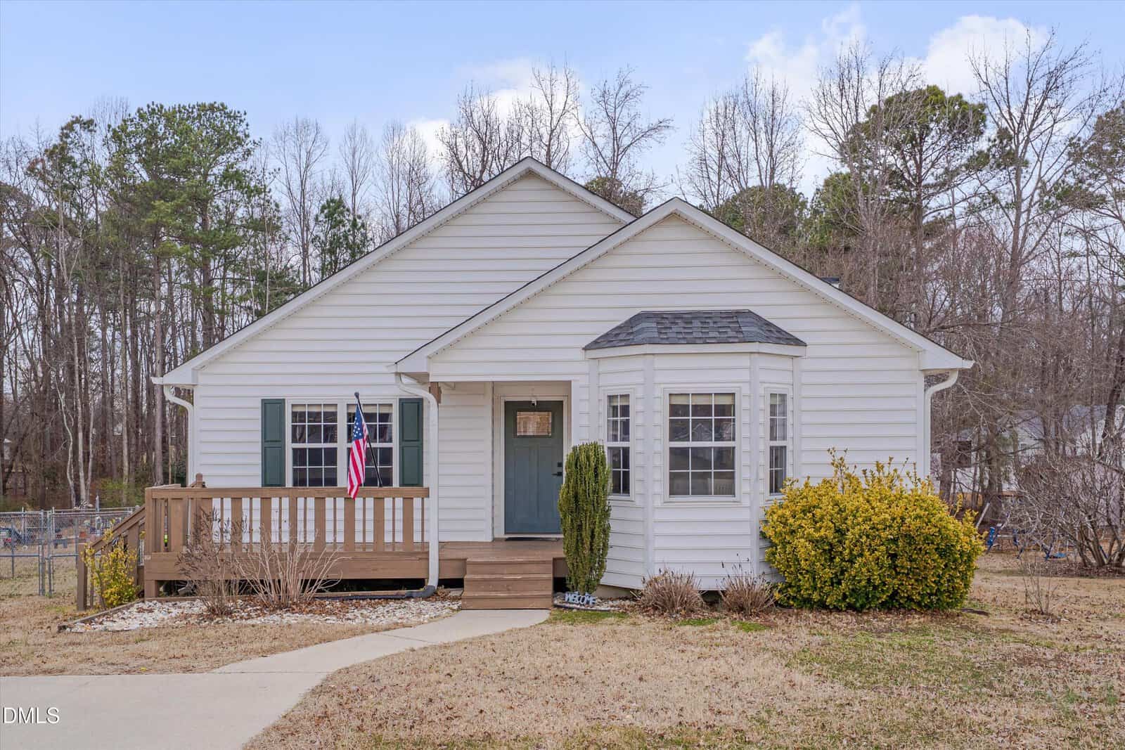 Charming single-story white house in Creedmoor with a green door, small porch, and an American flag. Shrubs line the front of this 304 Bridget Way residence, while leafless trees stand in the background. The yard features patchy grass and a curved concrete walkway for added character.