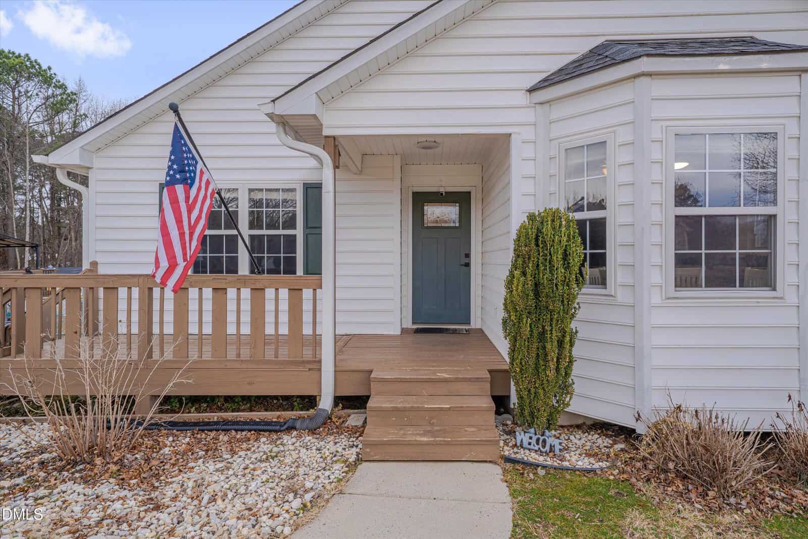 Single-story white house with a front porch, an American flag, a blue front door, and a small “Welcome” sign near a tall shrub. Rock landscaping lines the walkway leading up to the porch entrance.