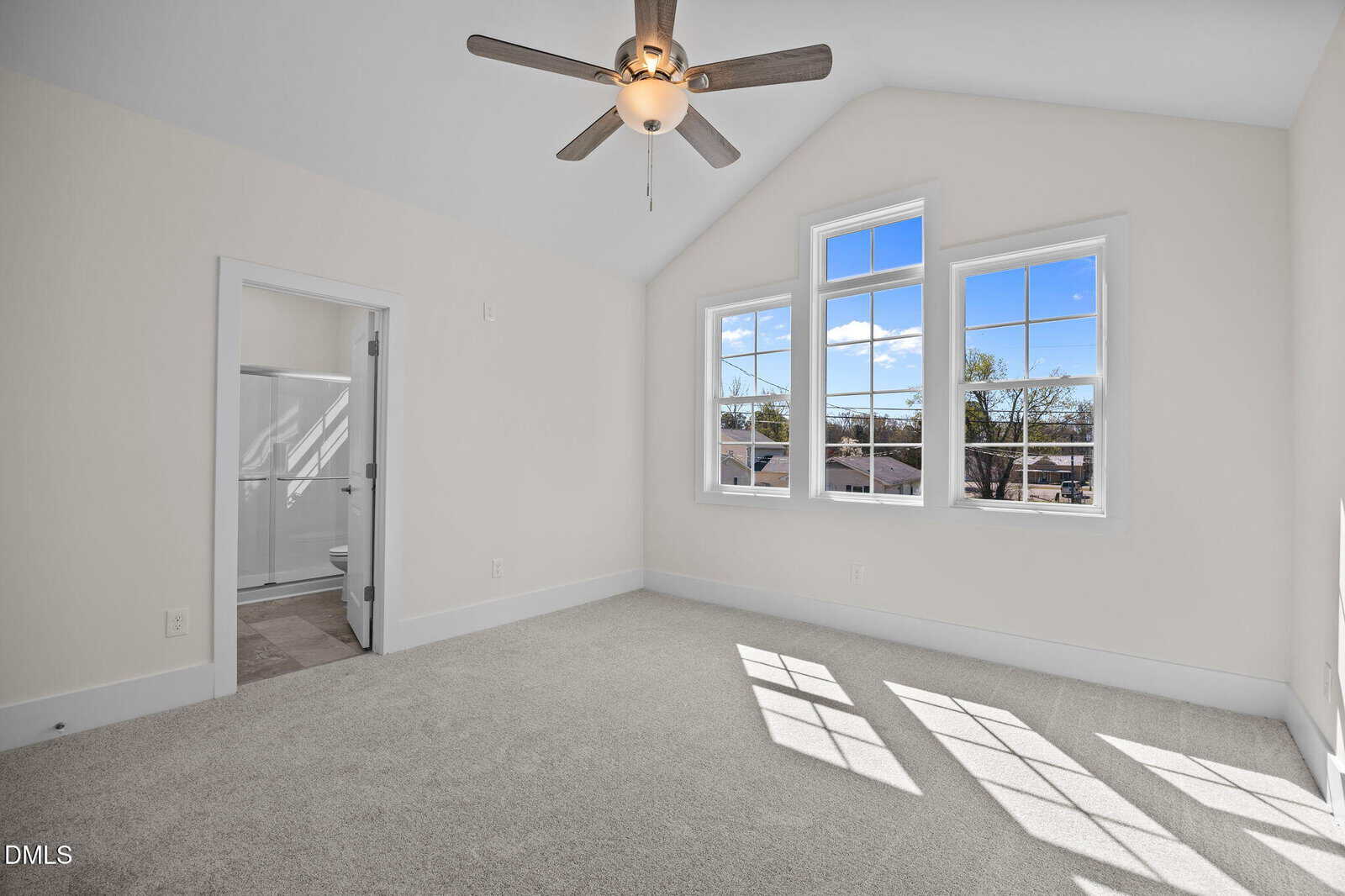 Bright, empty bedroom with beige carpet, white walls, a ceiling fan, large windows with sunlight, and an open door leading to a bathroom with a glass shower.