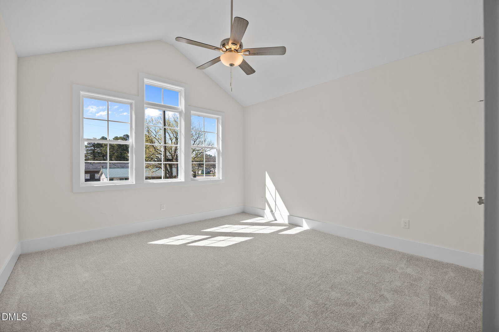 Empty white room with a vaulted ceiling, beige carpet, large windows letting in natural light, and a ceiling fan with lights. The view outside shows trees and a blue sky.