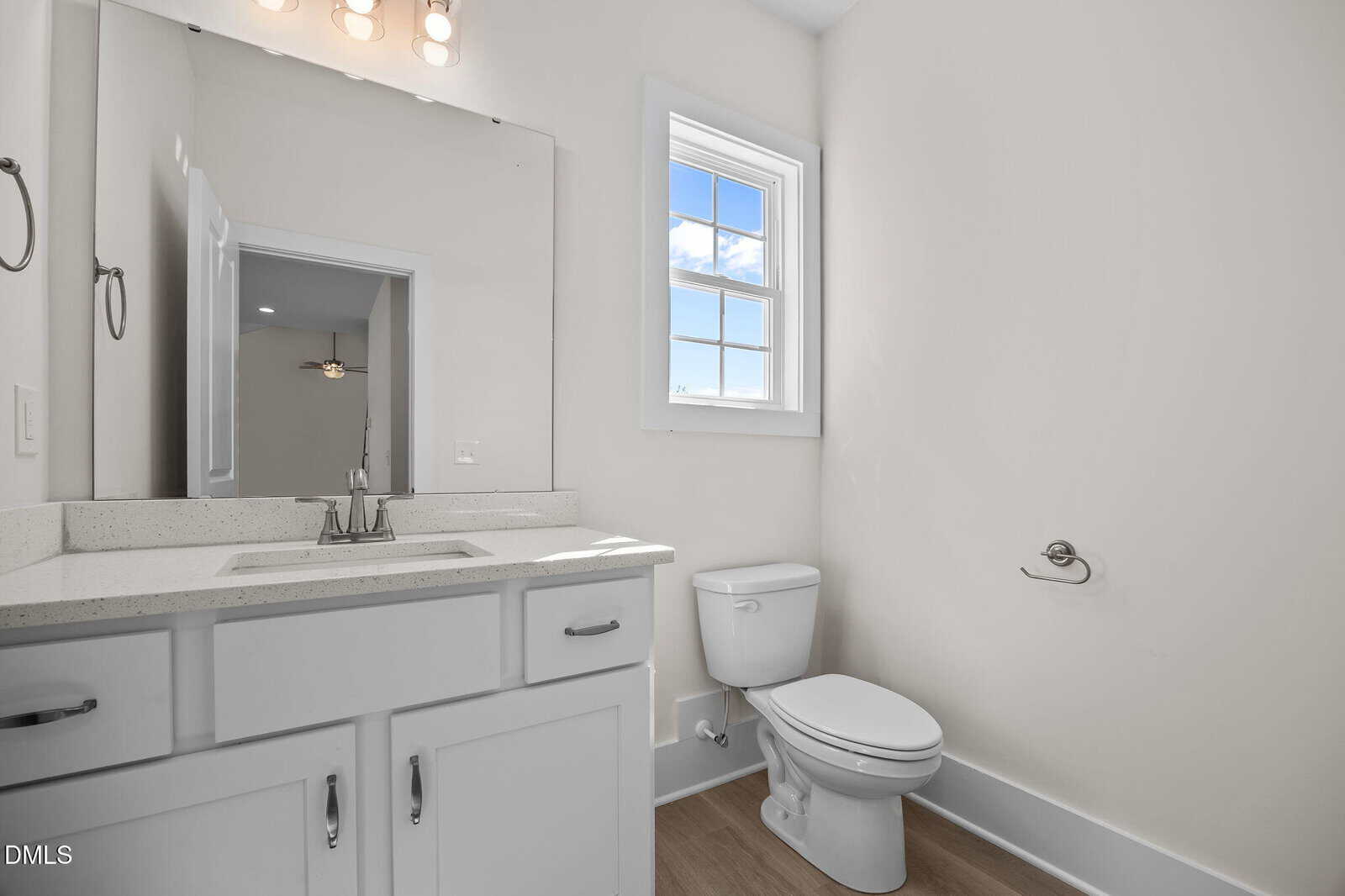 Modern bathroom with white walls, a large mirror above a white sink vanity, a window letting in natural light, a toilet, wood-look flooring, and a wall-mounted toilet paper holder.