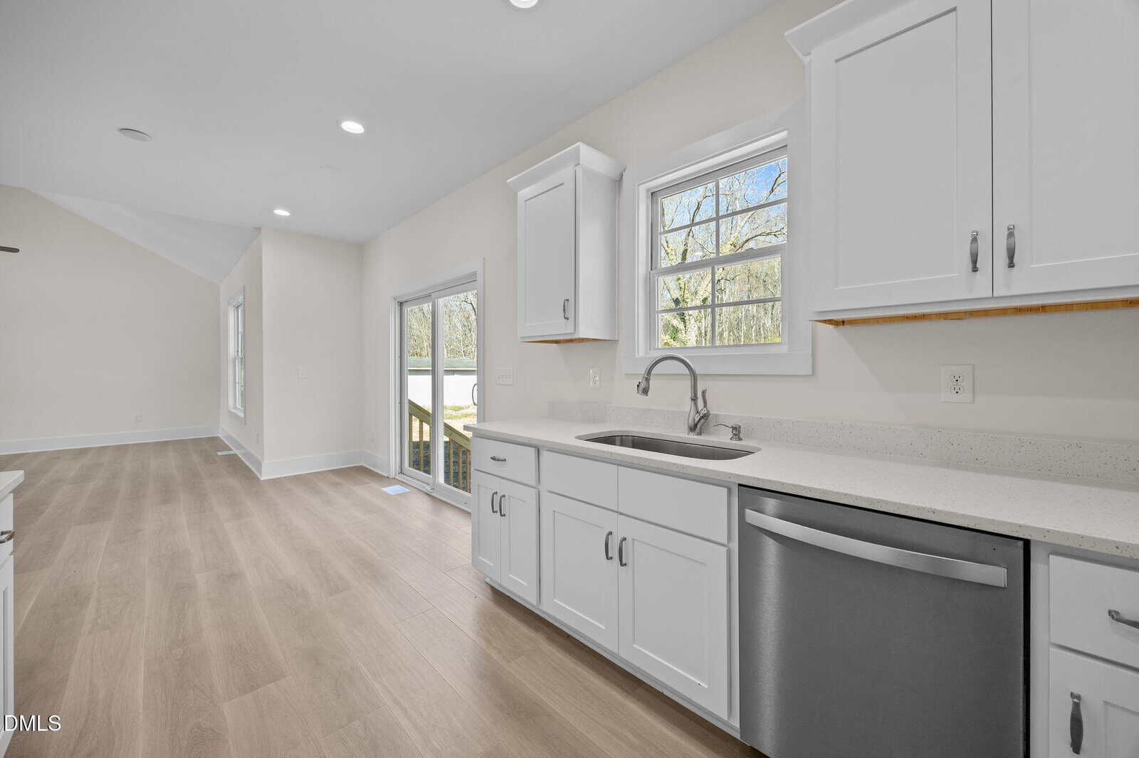 A modern kitchen with white cabinets, a stainless steel dishwasher, a sink under a window, and light wood flooring. Sliding glass doors open to an outdoor area, and the walls are painted light beige.