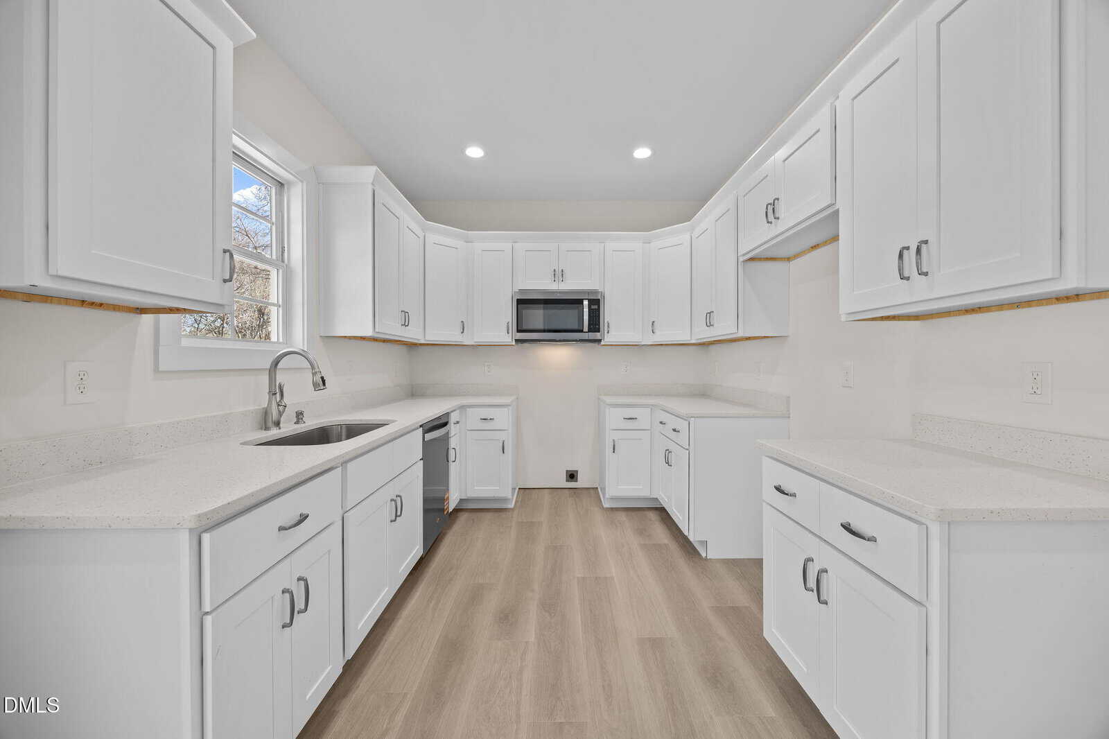 A modern kitchen with white cabinets, white countertops, a stainless steel sink under a window, a built-in microwave, and light wood-style flooring. The room is well-lit with ceiling lights.