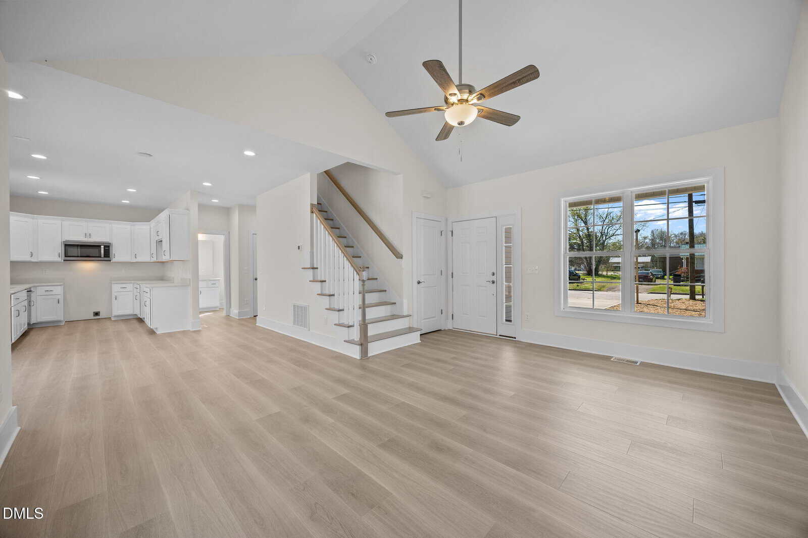 Open-concept living area with light wood flooring, white walls, and a ceiling fan. A staircase leads upstairs, and a large window brings in natural light. The kitchen is visible in the background with white cabinets.