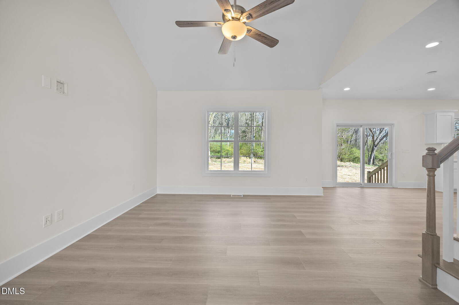 A spacious, empty room with light wood flooring, white walls, a ceiling fan with a light, two large windows, and a partial view of a staircase and an open doorway leading outside.