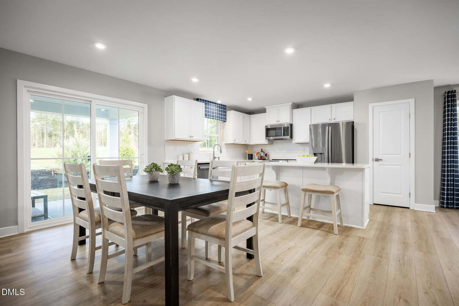 Modern kitchen and dining area with wood flooring, white cabinets, stainless steel appliances, a black dining table with six chairs, and a breakfast bar with two stools. Large window and sliding glass door let in natural light.