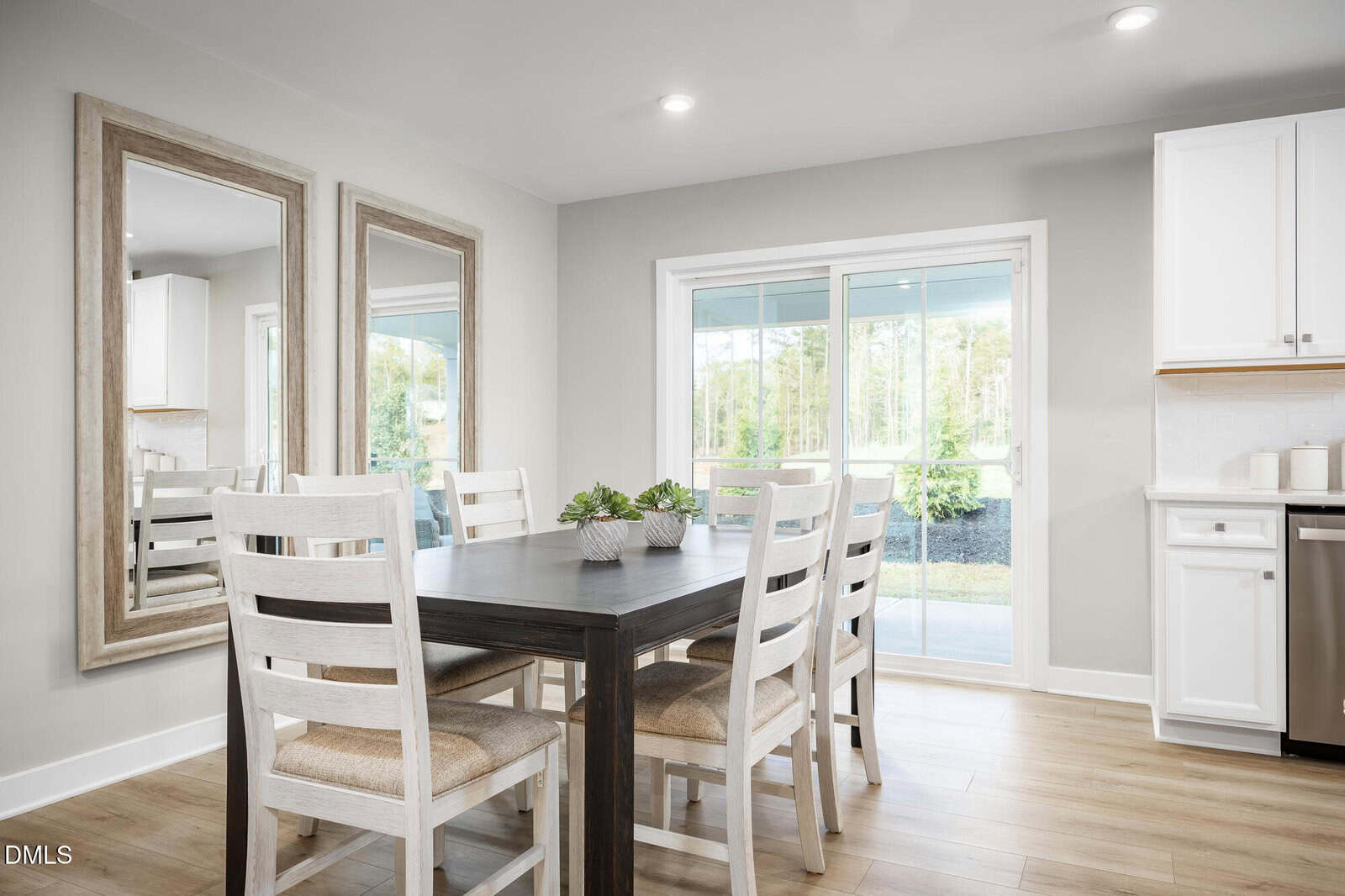 A dining area with a dark wooden table, four white chairs, two large mirrors on the wall, sliding glass doors leading outside, and white kitchen cabinets on the right. A small plant sits on the table.