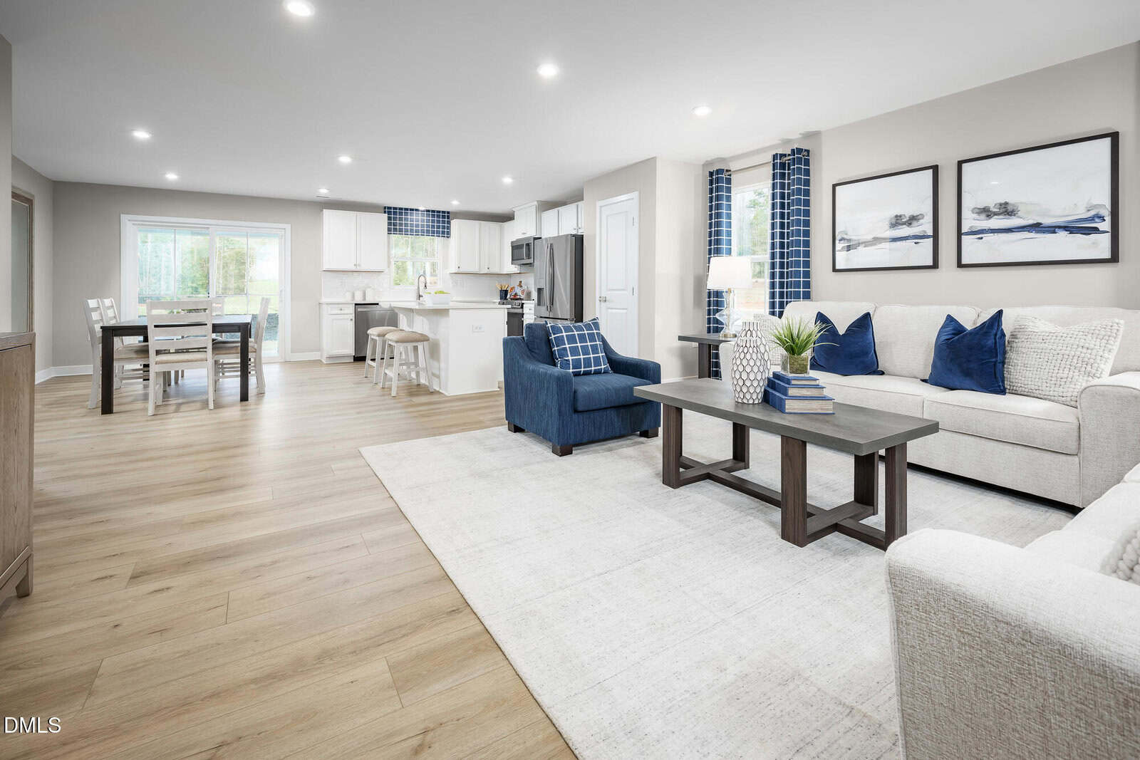 Open-concept living and dining area with light wood flooring, neutral walls, blue and white furniture, modern kitchen in the background, large windows, and recessed lighting.