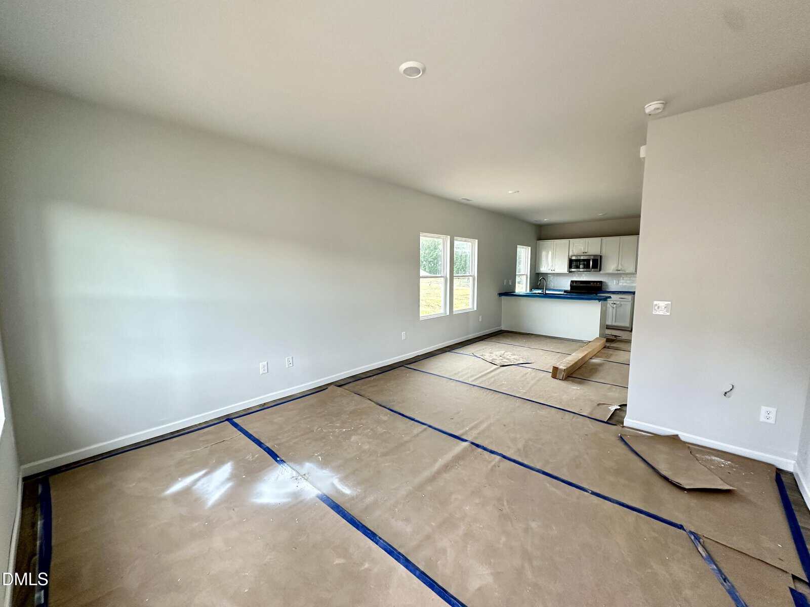 Unfurnished room with protected floors, gray walls, large windows, and an open kitchen area with white cabinets and stainless steel appliances in the background.