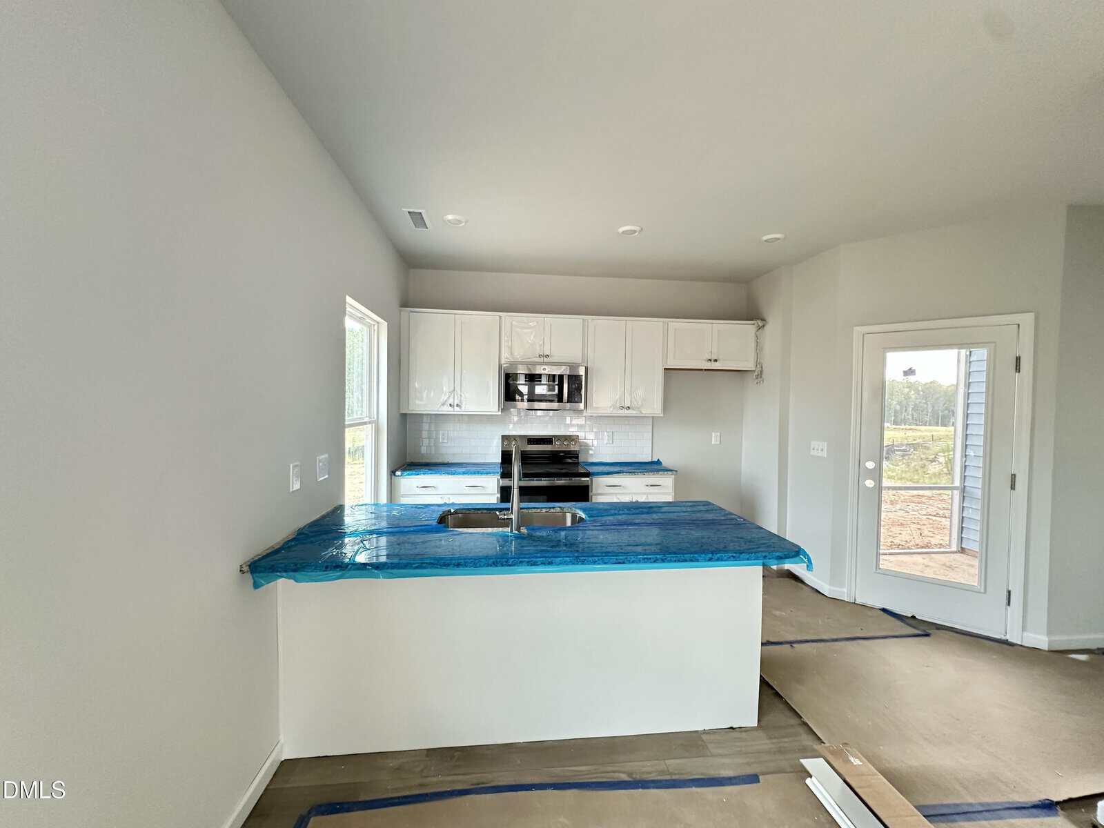 A newly constructed kitchen with white cabinets, a center island covered in blue protective wrap, stainless steel appliances, and a door leading outside. The floor is partially covered with protective material.