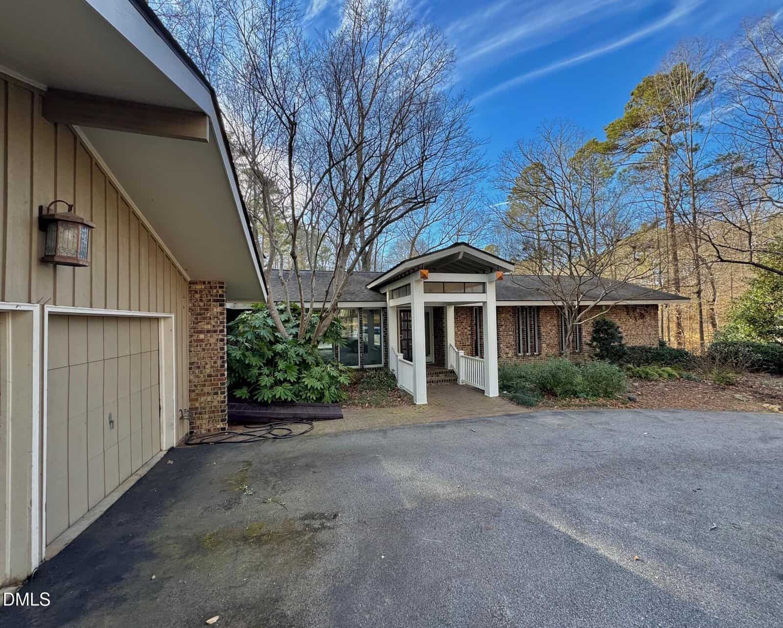 A single-story brick house with a covered entryway, white columns, and a two-car garage on the left. Leafless trees and green shrubs surround the house under a blue sky with wispy clouds.