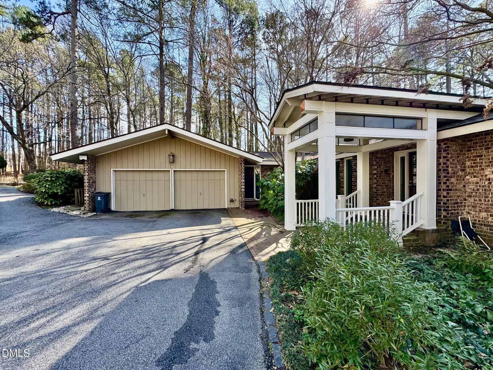 A suburban home with a brick exterior, covered front porch, white columns, and a detached two-car garage. Tall trees and greenery surround the house, and sunlight filters through the branches.