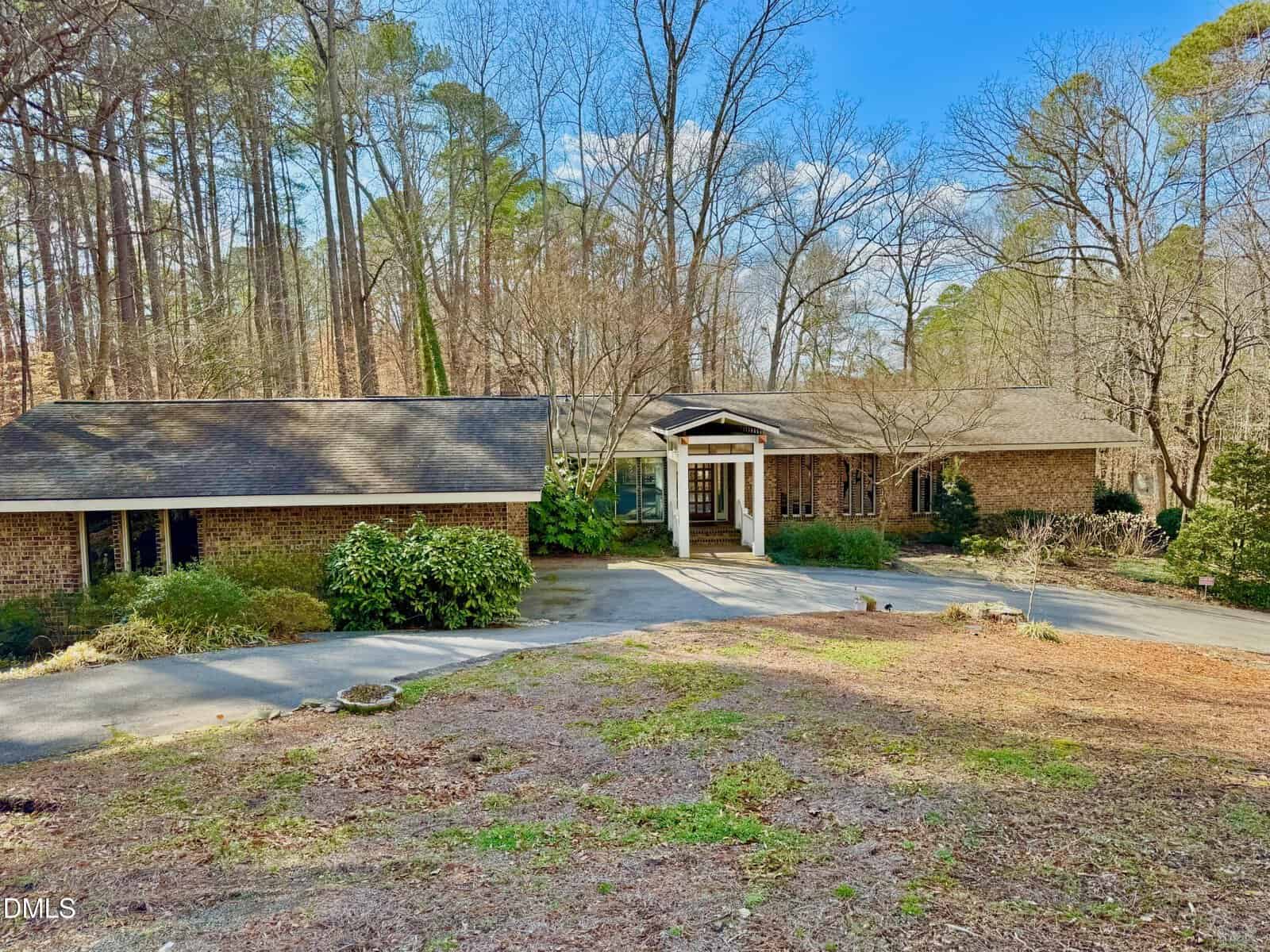 A single-story brick house with a covered entryway, set among tall trees. The driveway curves in front, and there is sparse landscaping with some bushes and bare trees. The sky is blue with a few clouds.