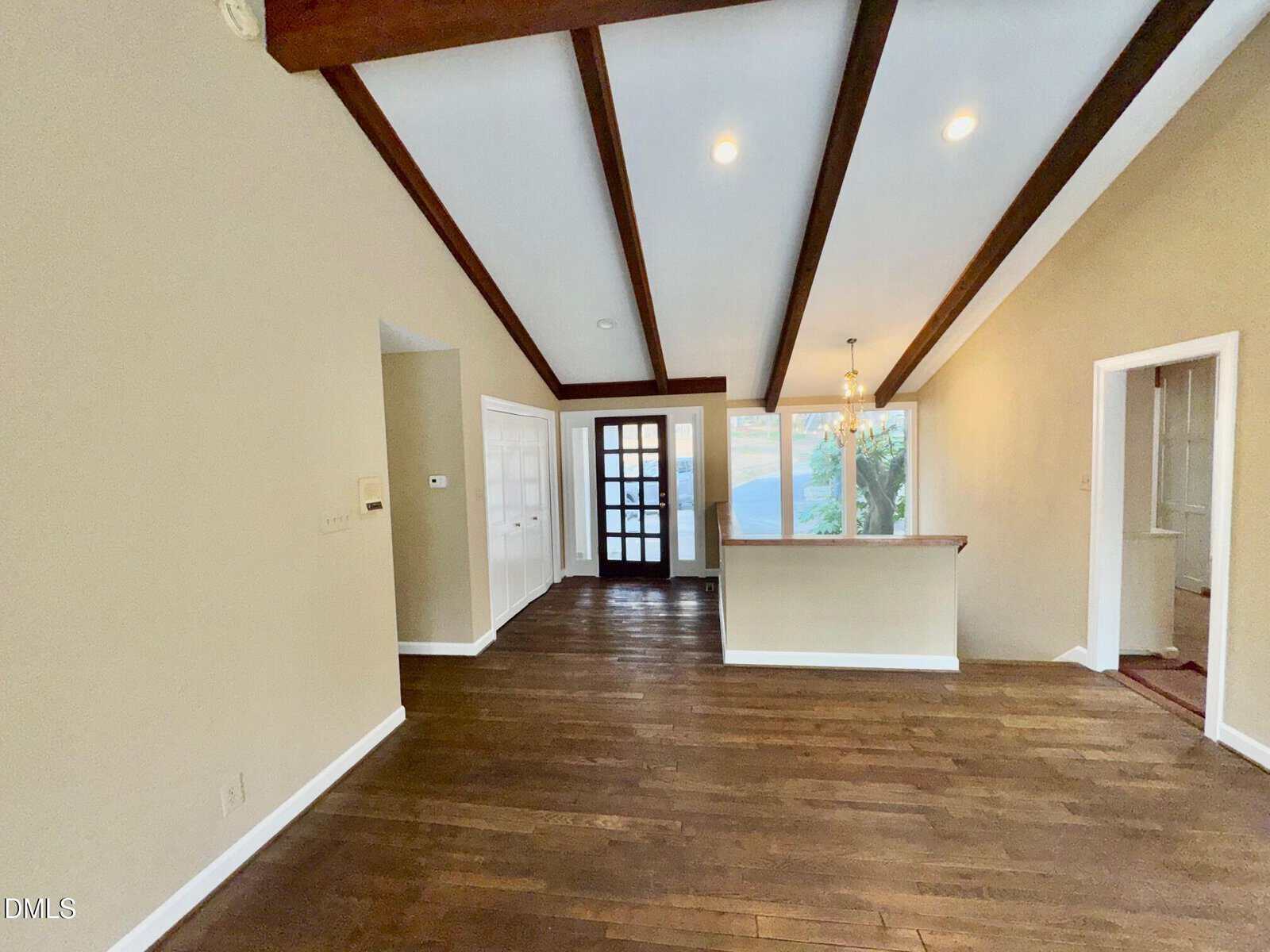 Open living room with wood floors, exposed ceiling beams, beige walls, and a view of the entry door, dining area, and kitchen pass-through window. Large windows provide natural light.