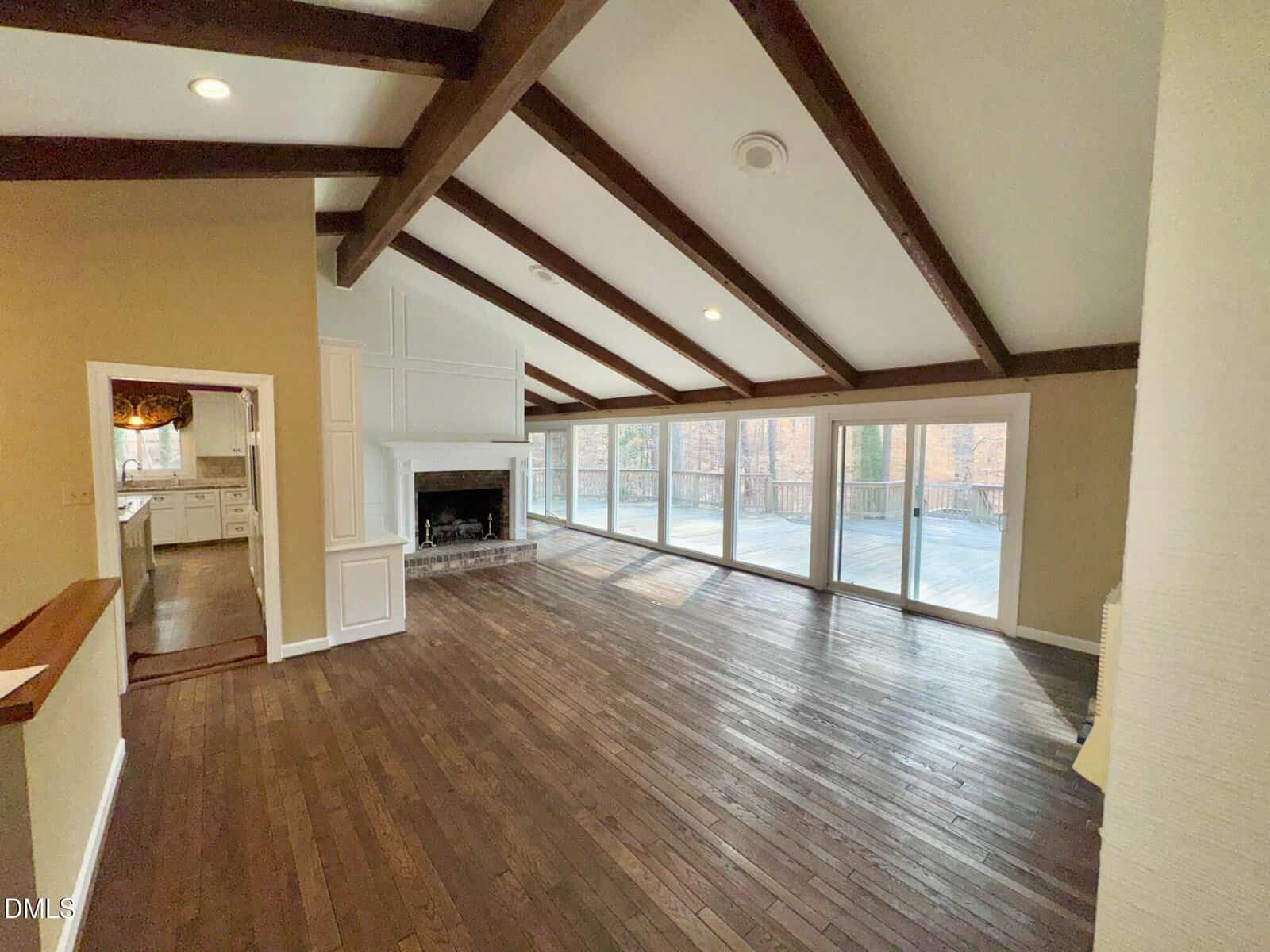 Spacious living room with vaulted ceiling, exposed wooden beams, fireplace, large windows, and sliding glass doors opening to a deck. Hardwood floors throughout. Kitchen visible through doorway on the left.