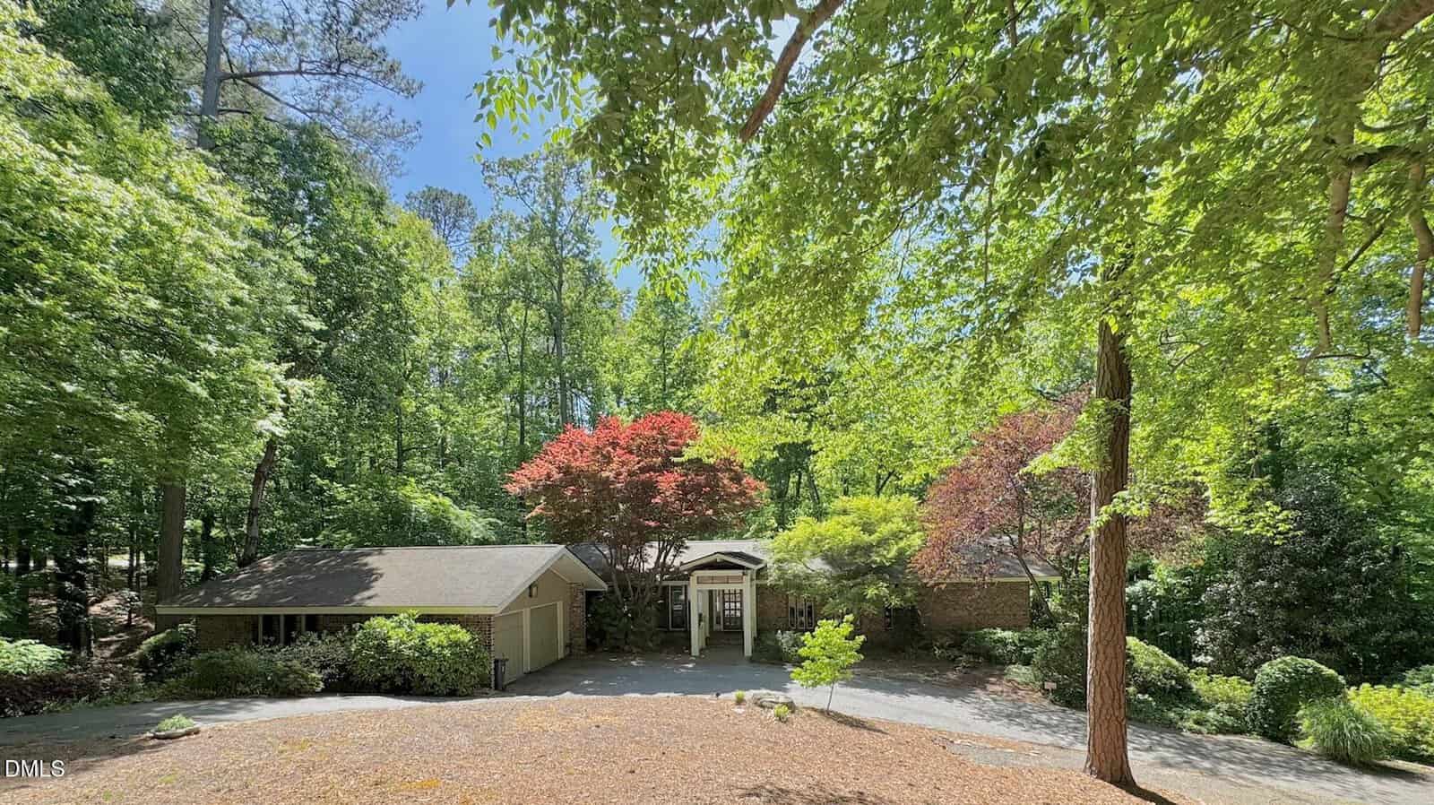A single-story house with a sloped roof sits among tall green trees and colorful foliage. Sunlight filters through the leaves, casting shadows on the driveway and front yard. The setting appears calm and secluded.
