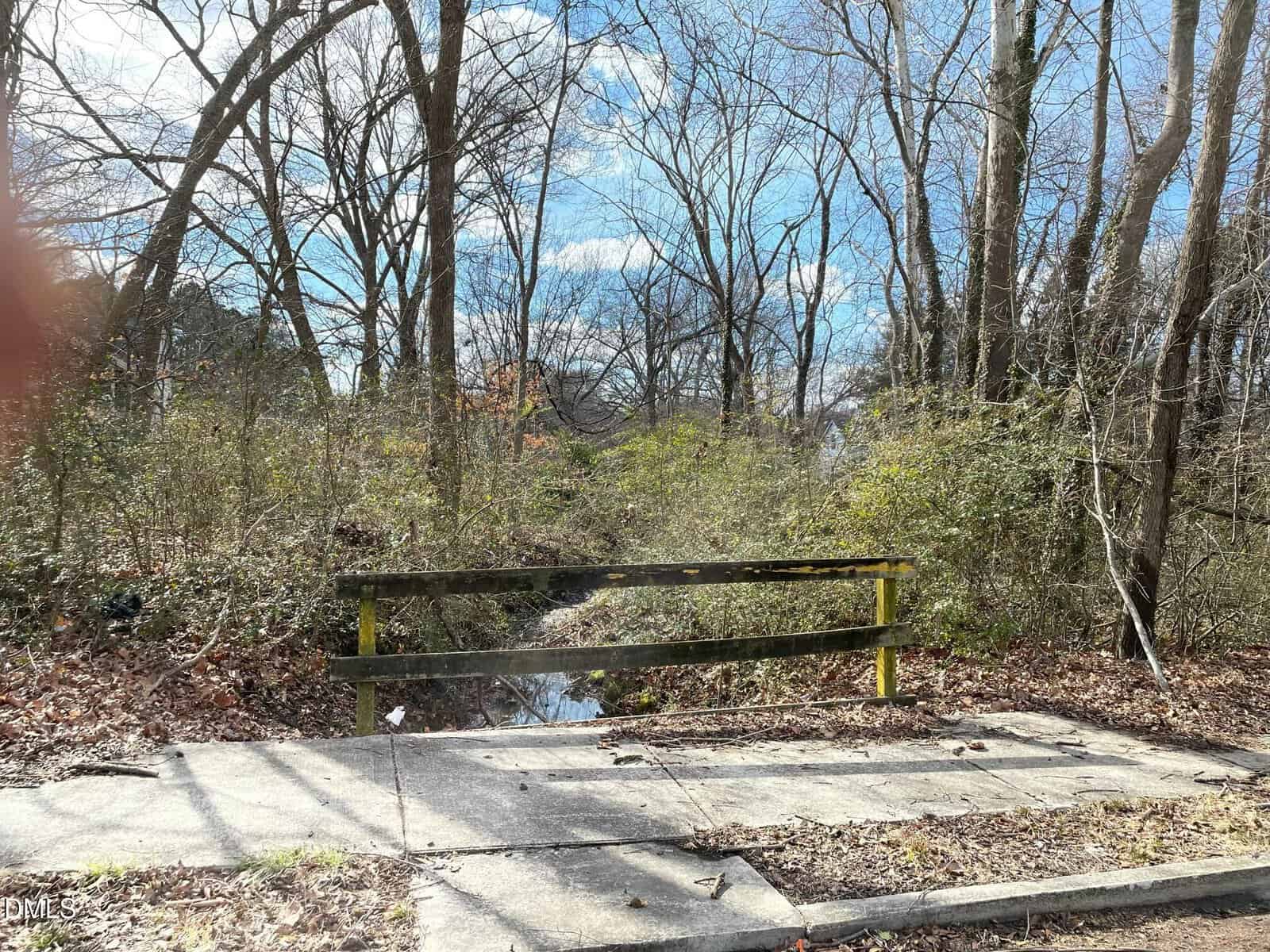 A small wooden guardrail sits at the edge of a concrete sidewalk, overlooking a narrow creek surrounded by leafless trees and dense underbrush, under a partly cloudy sky.