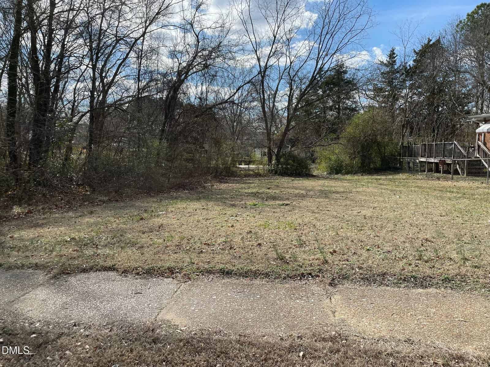 A mostly empty lot with short grass and scattered bushes, bordered by trees and a sidewalk in the foreground. A wooden porch is visible on the right, and the sky is partly cloudy.