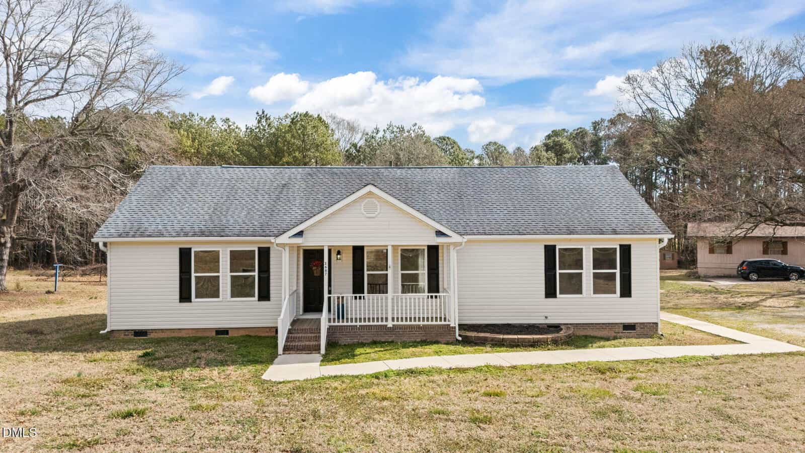 Single-story house with white siding, black shutters, and a covered front porch located at 2687 NC 39 S, Henderson. The home sits on a grassy lot with a concrete walkway, surrounded by trees and neighboring houses under a partly cloudy sky.