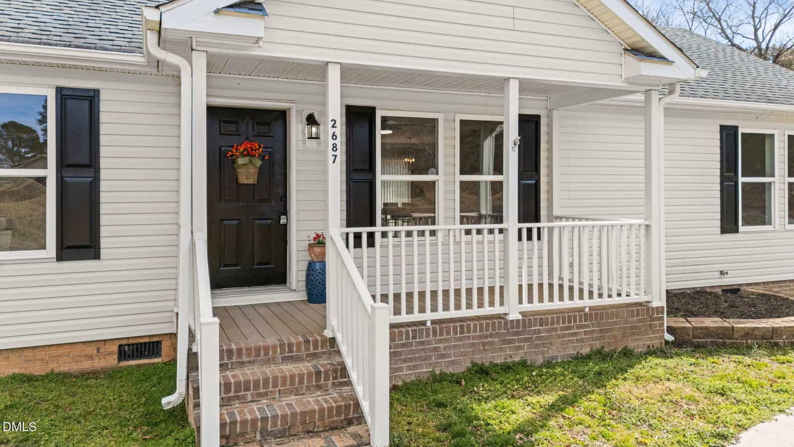 A small front porch with white railings and steps leads to a black door with a flower basket. The house has white siding, brick base, and visible windows. House number 2687 is displayed next to the door.