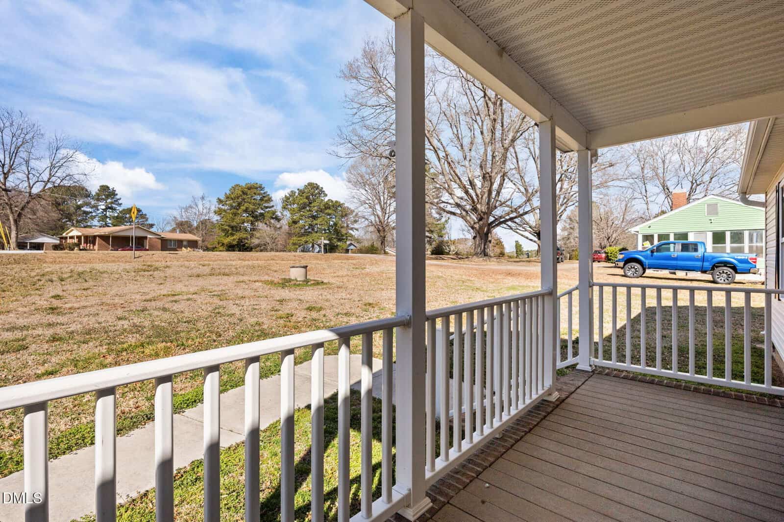 View from a covered porch with white railings overlooking a grassy yard, a sidewalk, and a residential street with houses and a blue pickup truck parked on the right. Trees are scattered throughout the background.