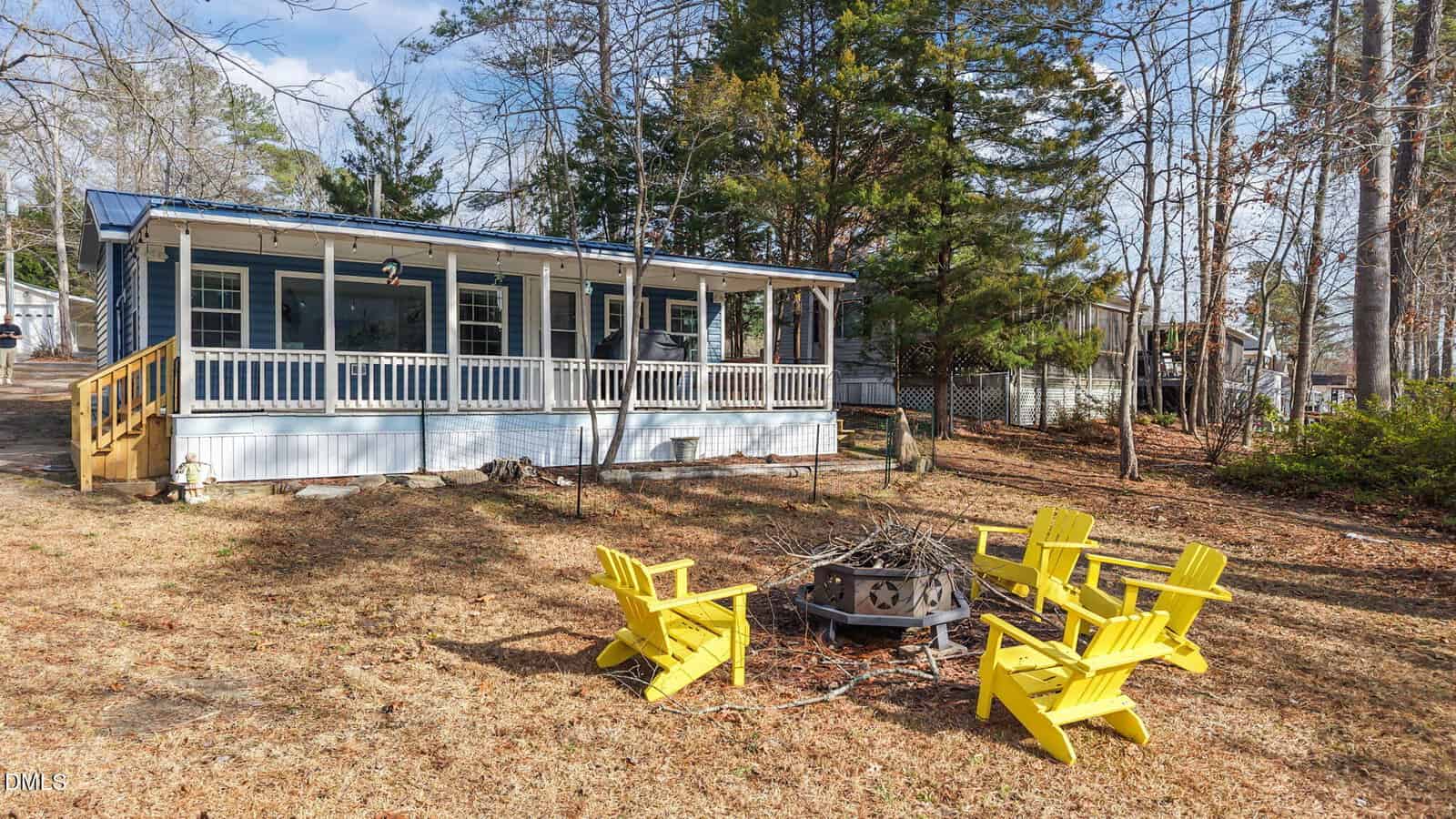 A blue house with a large front porch sits among trees. In the yard, four bright yellow Adirondack chairs are arranged around a fire pit on dry grass. The sky is partly cloudy.