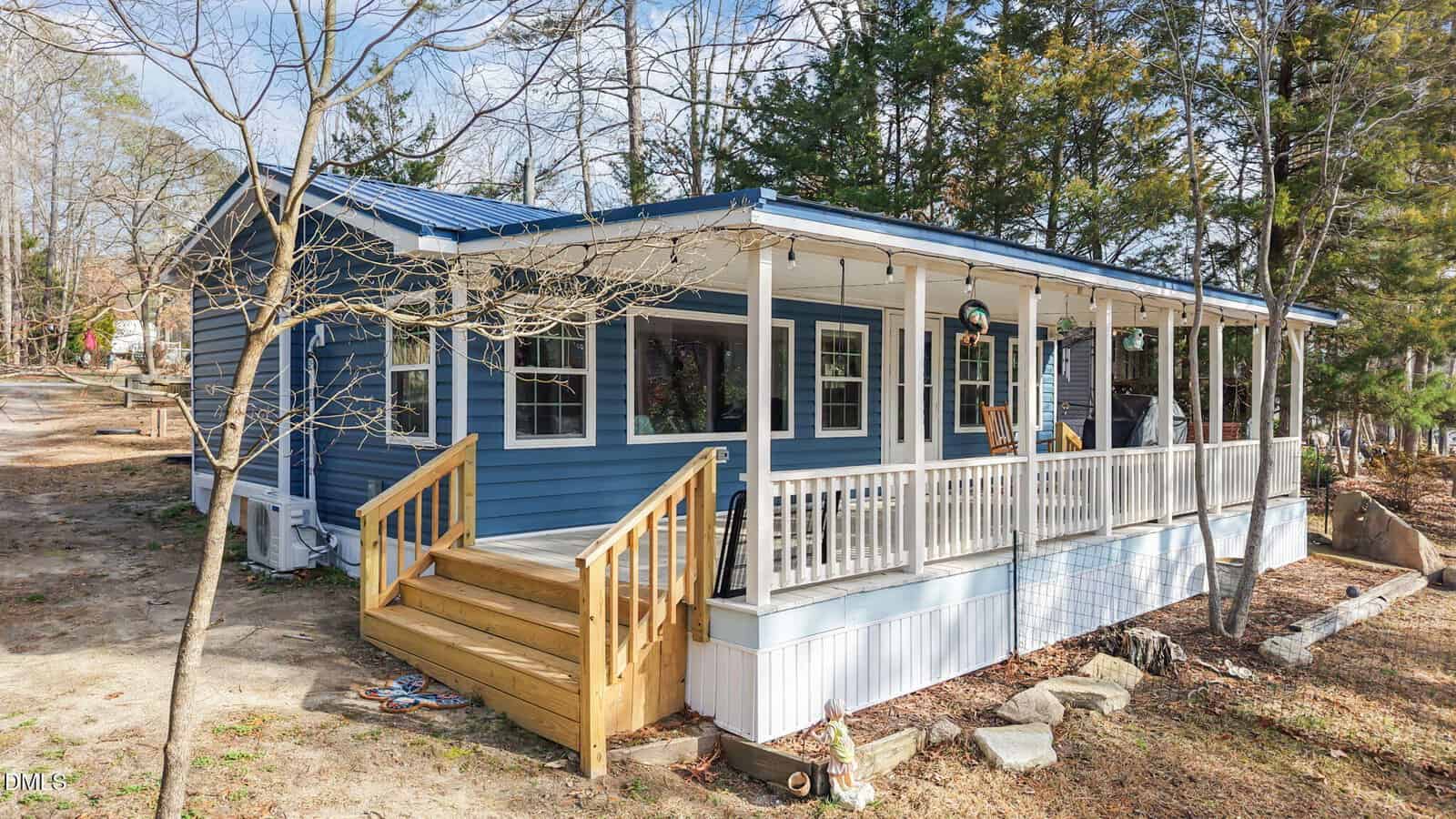 A blue house with white trim and a covered front porch, surrounded by trees. Wooden steps lead up to the porch, which features white railings. The yard is partly bare soil with scattered rocks and leaves.