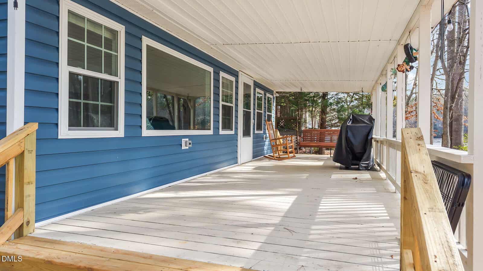 A spacious front porch with light wooden flooring, blue siding, several large windows, a wooden rocking chair, a porch swing, a covered grill, and a wooden railing, overlooking a wooded area.