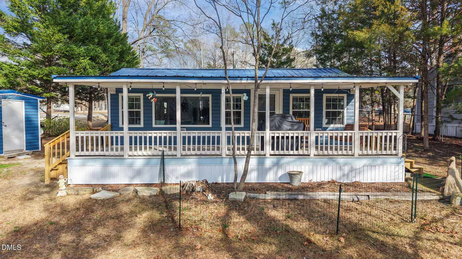 A blue and white house with a large covered front porch, white railings, and a metal roof, surrounded by trees and a fenced yard. There is outdoor furniture and a grill on the porch.