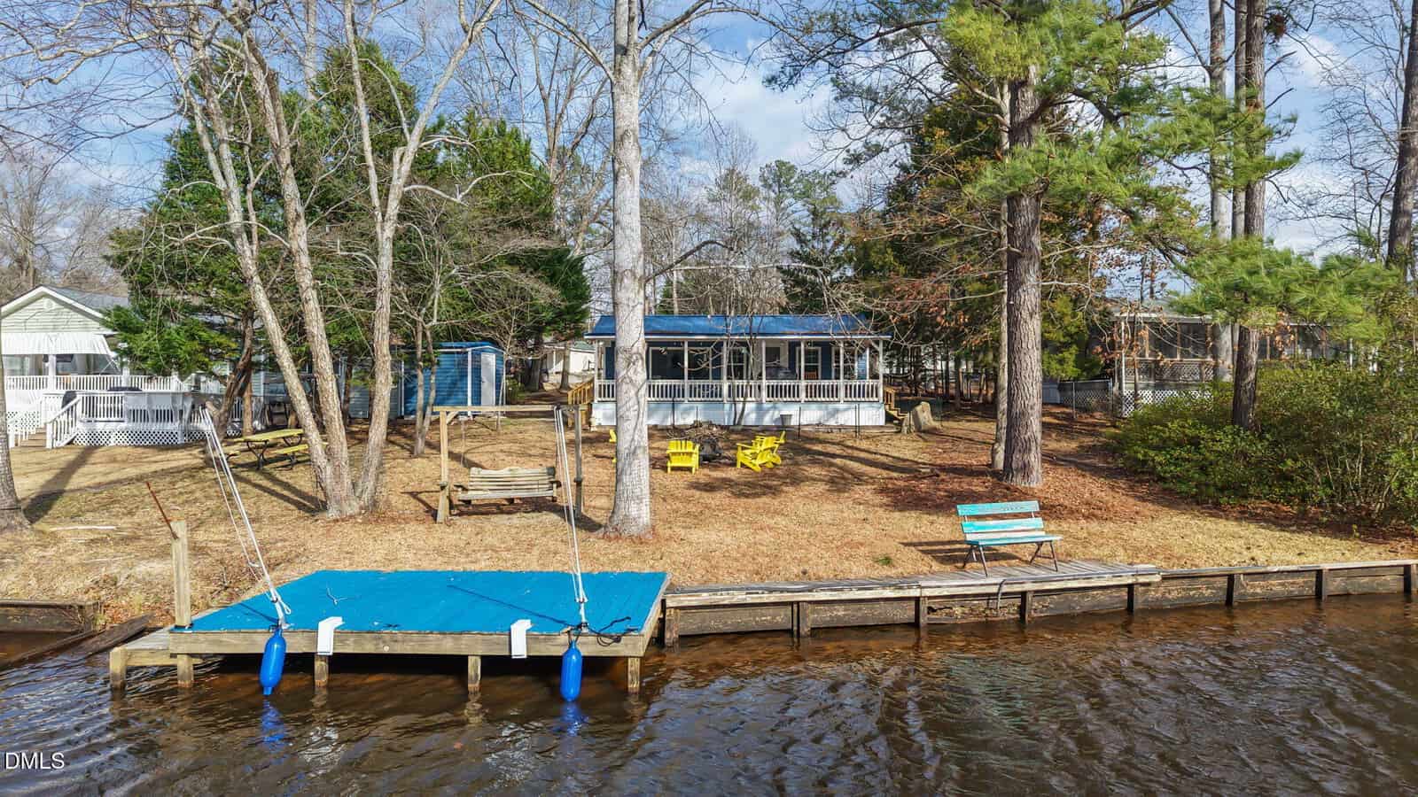 A small lakeside cabin at 1607 Sagamore Drive, Louisburg, with a screened porch, sits among tall trees. In front are yellow Adirondack chairs, a wooden swing, and a blue bench. A dock with a blue cover stretches into the water.
