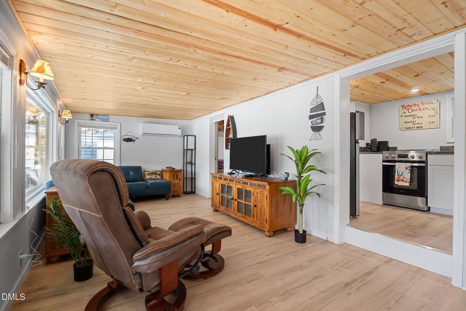 Cozy living room with wood ceiling and floors, brown recliner, blue sofa, TV on wooden stand, plants, and open view into a white kitchen with stainless steel appliances and a hand-written sign above the stove.