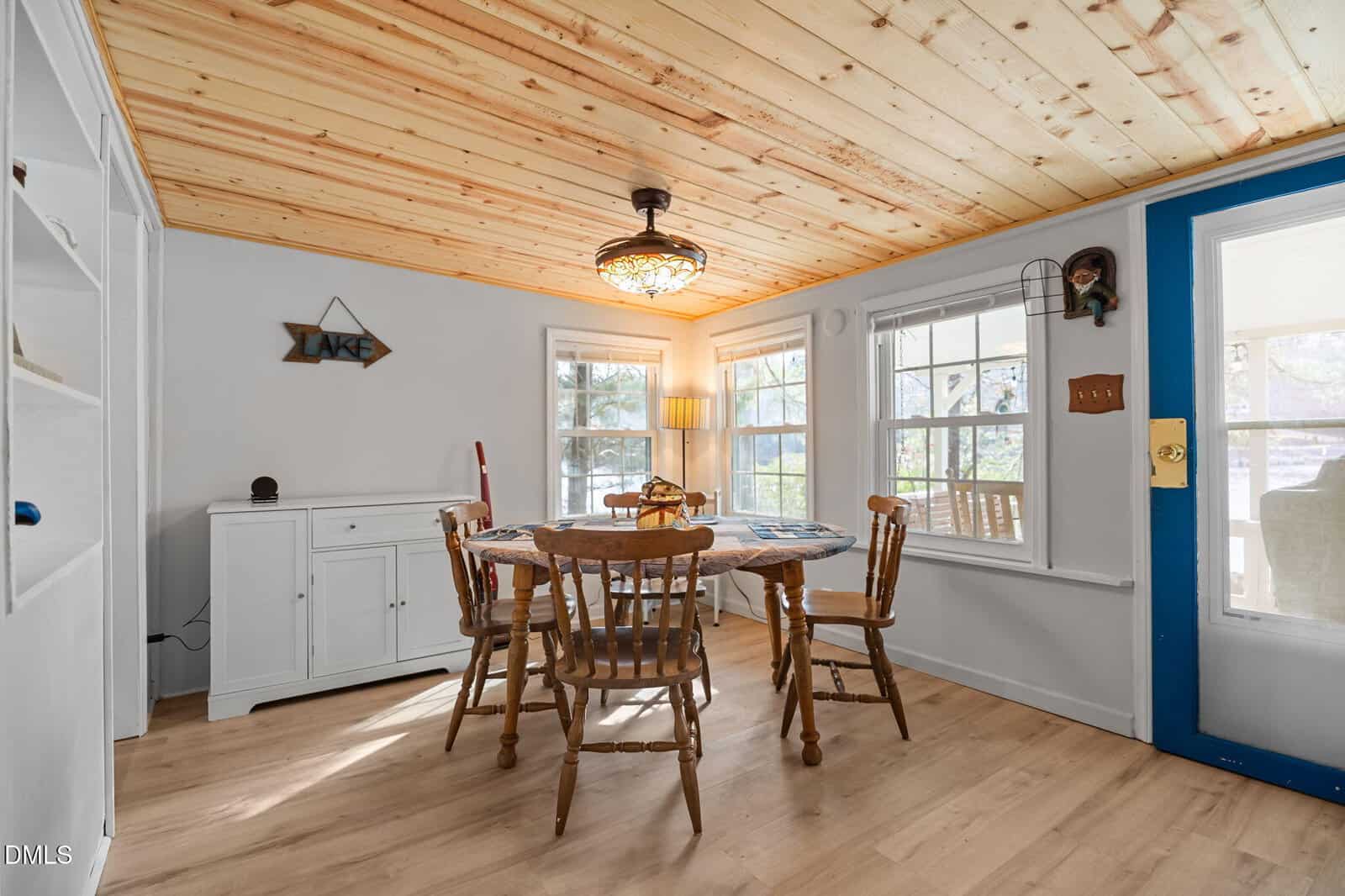 A cozy dining room with a wooden table and four chairs, white walls, a wood ceiling, lots of natural light from multiple windows, and simple decor including a “LAKE” sign.