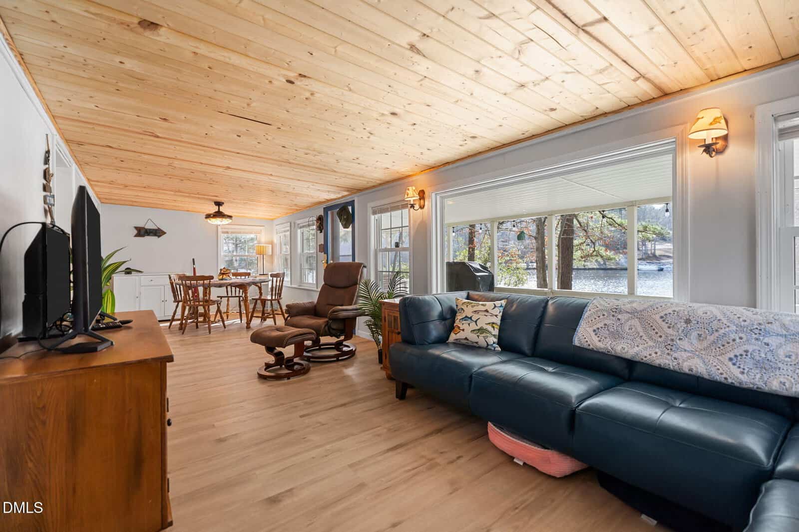 Bright sunroom with wooden ceiling, large windows, blue sectional sofa, recliner, TV on a stand, and dining table set near the back. Trees and water are visible outside the windows.