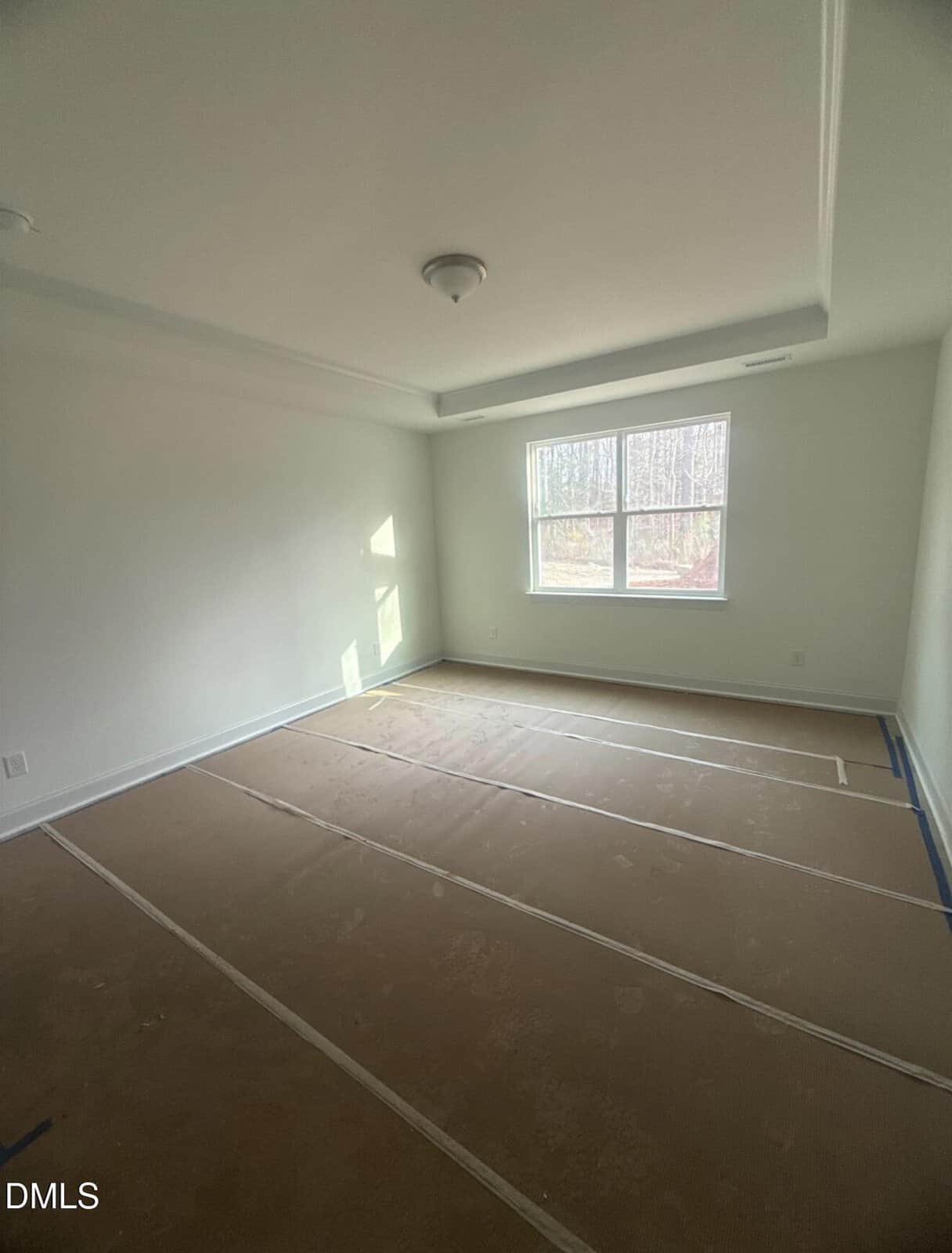Empty room with white walls, a ceiling light fixture, a double window letting in sunlight, and floors covered with brown protective paper secured by tape. No furniture or decorations are present.